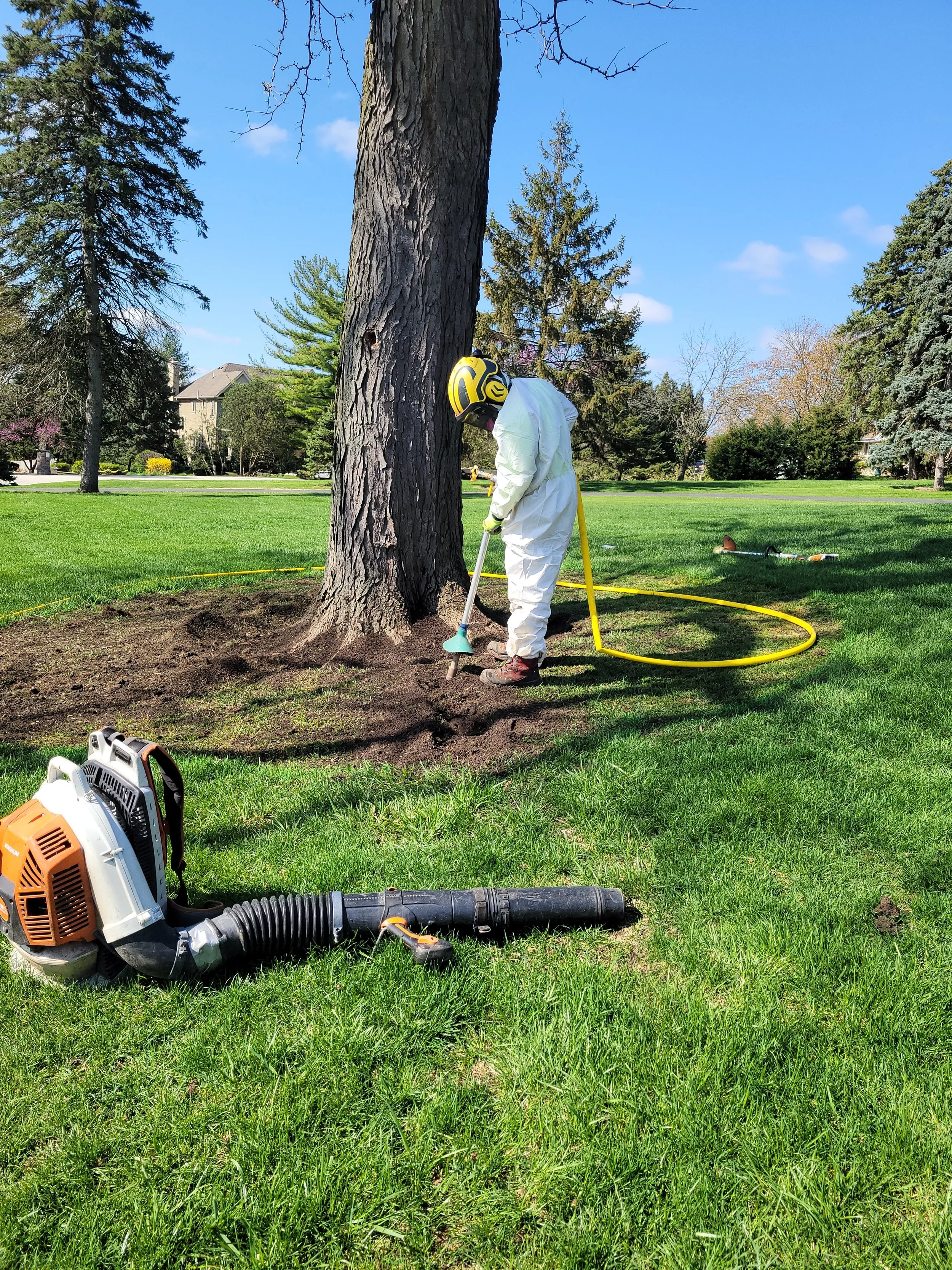 A person wearing protective gear including a helmet, gloves, and a suit is removing a tree stump from the ground using a tool, with a leaf blower or vacuum nearby on the grass.