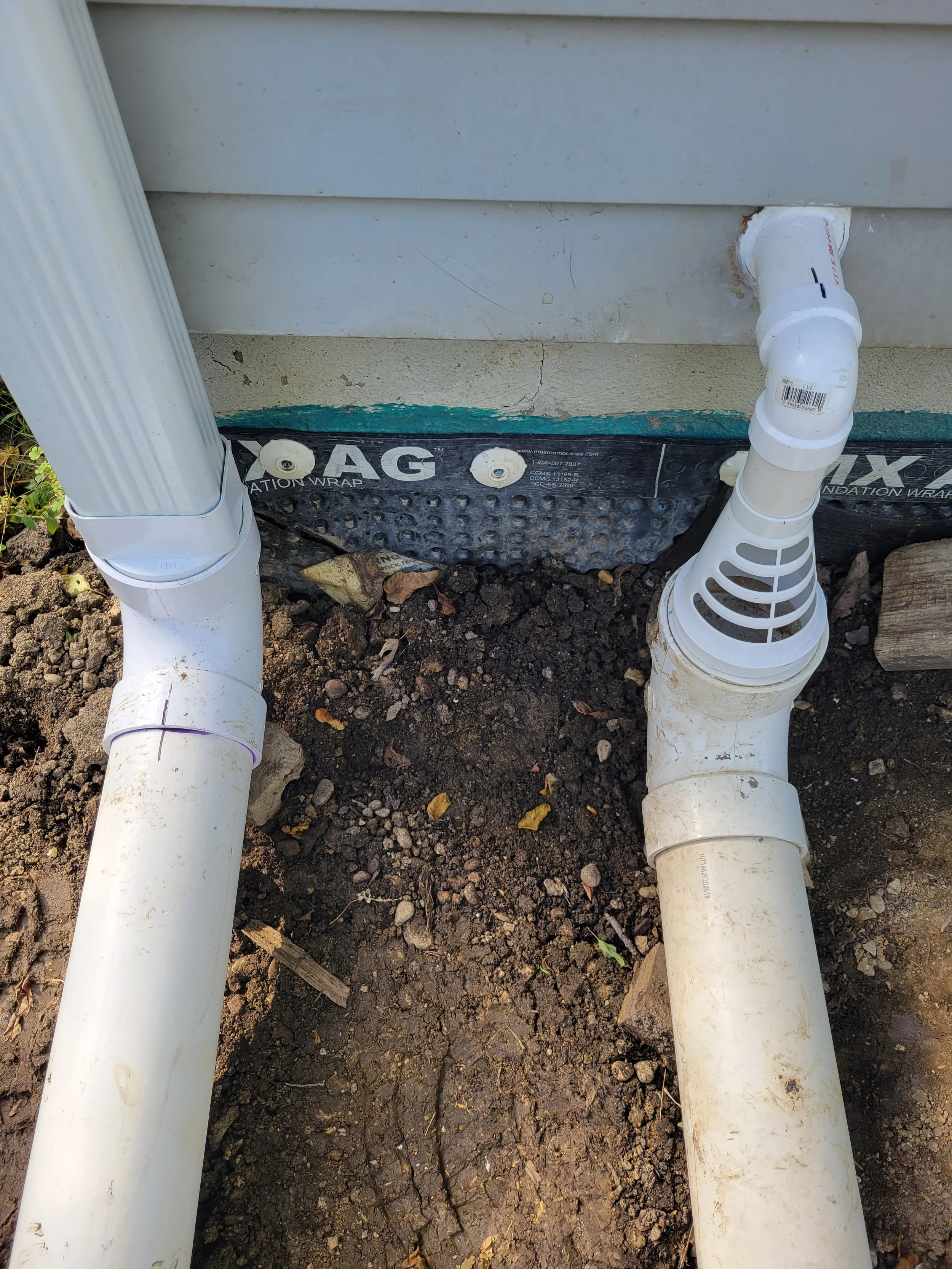 Underground plumbing pipes connected to a building foundation, with soil and dirt surrounding them, and a section of the building wall visible.