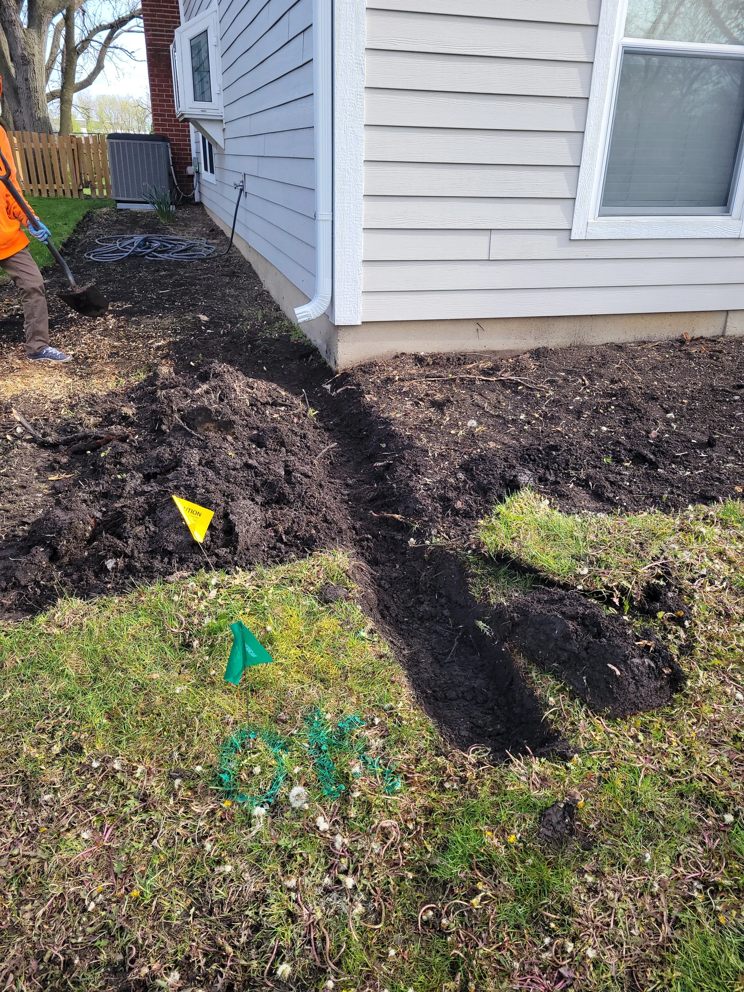 Backyard garden area with freshly dug trench along the foundation of a house, a worker in orange shirt and brown pants shoveling dirt, and small green and yellow flags marking spots in the soil.