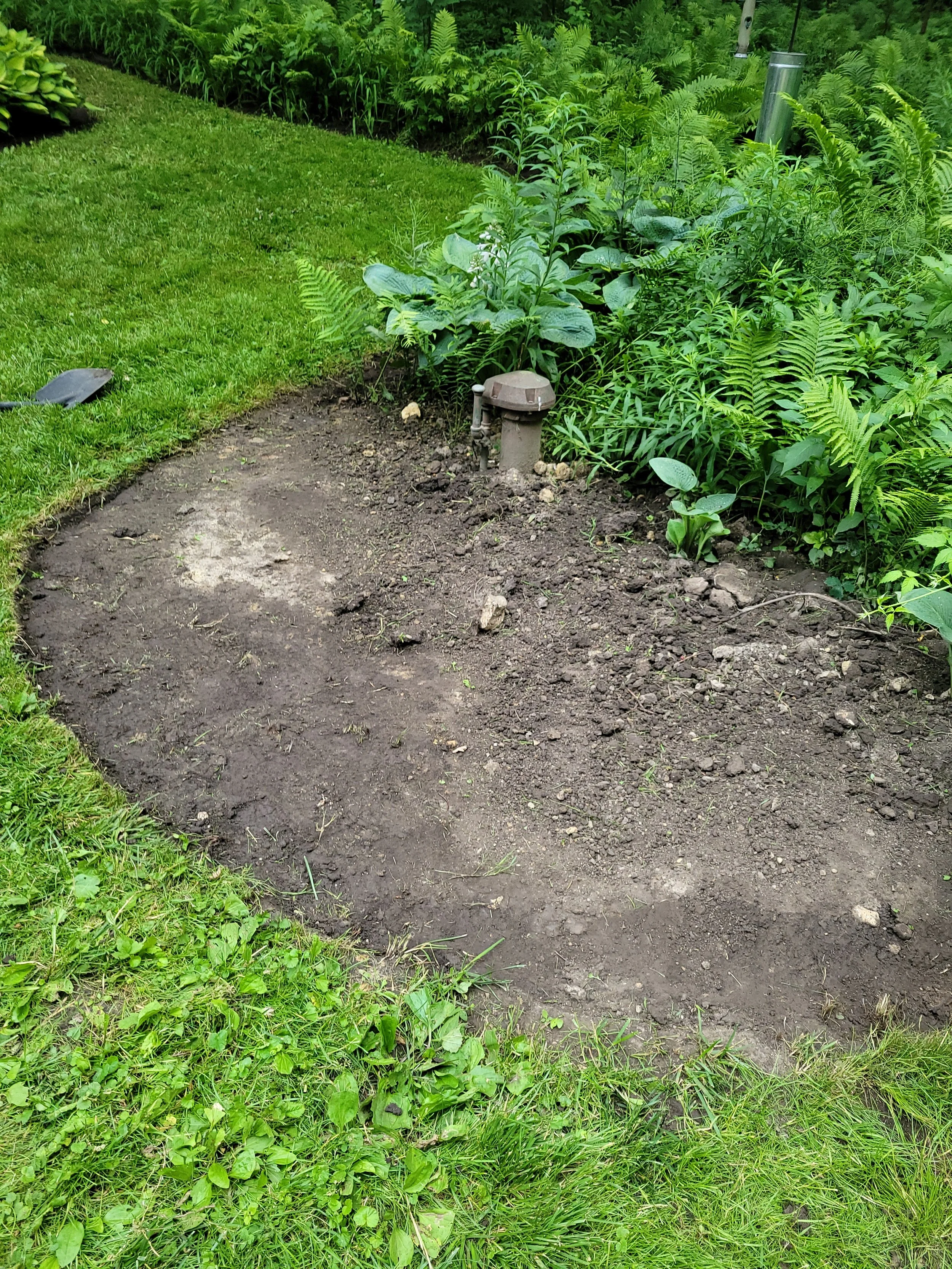 A newly dug garden bed with dirt, surrounded by green grass and plants, and a small outdoor lamp fixture in the middle.