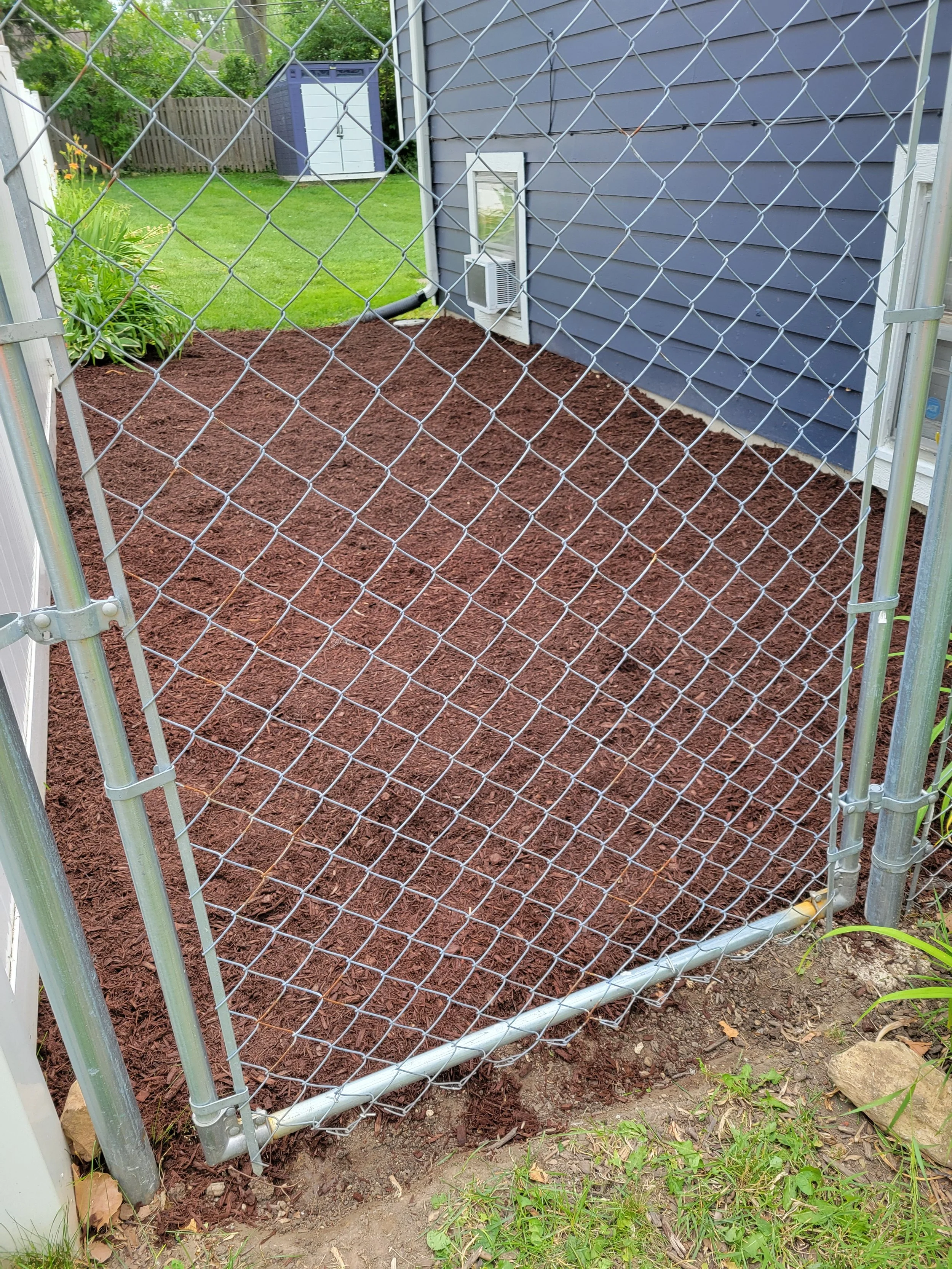 A newly prepared garden bed with reddish-brown soil, enclosed by a metal chain-link fence, next to a blue house with an air conditioning unit in a backyard.