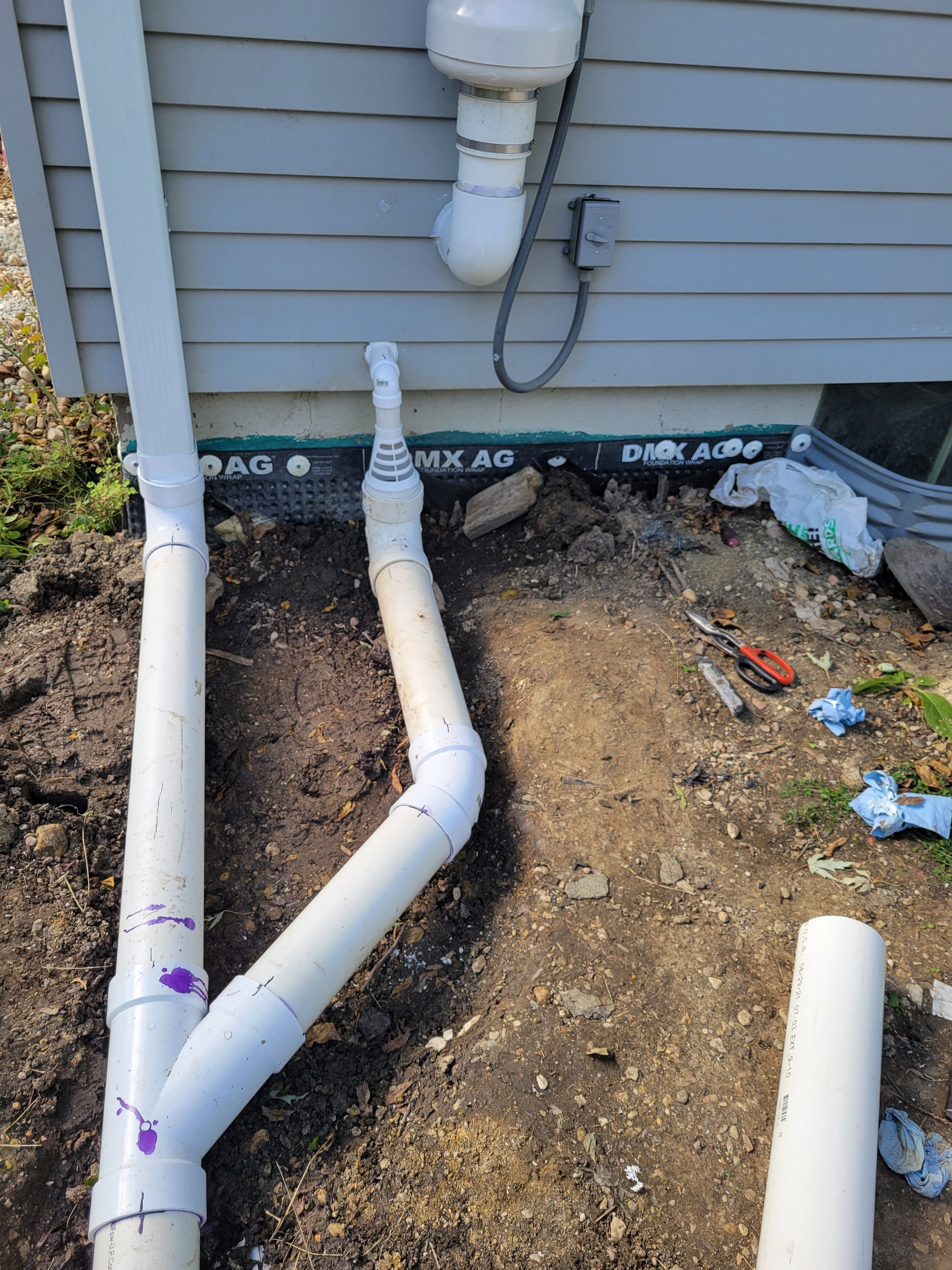 Underground plumbing pipes being installed outside a house with gray siding, with tools and construction materials around.