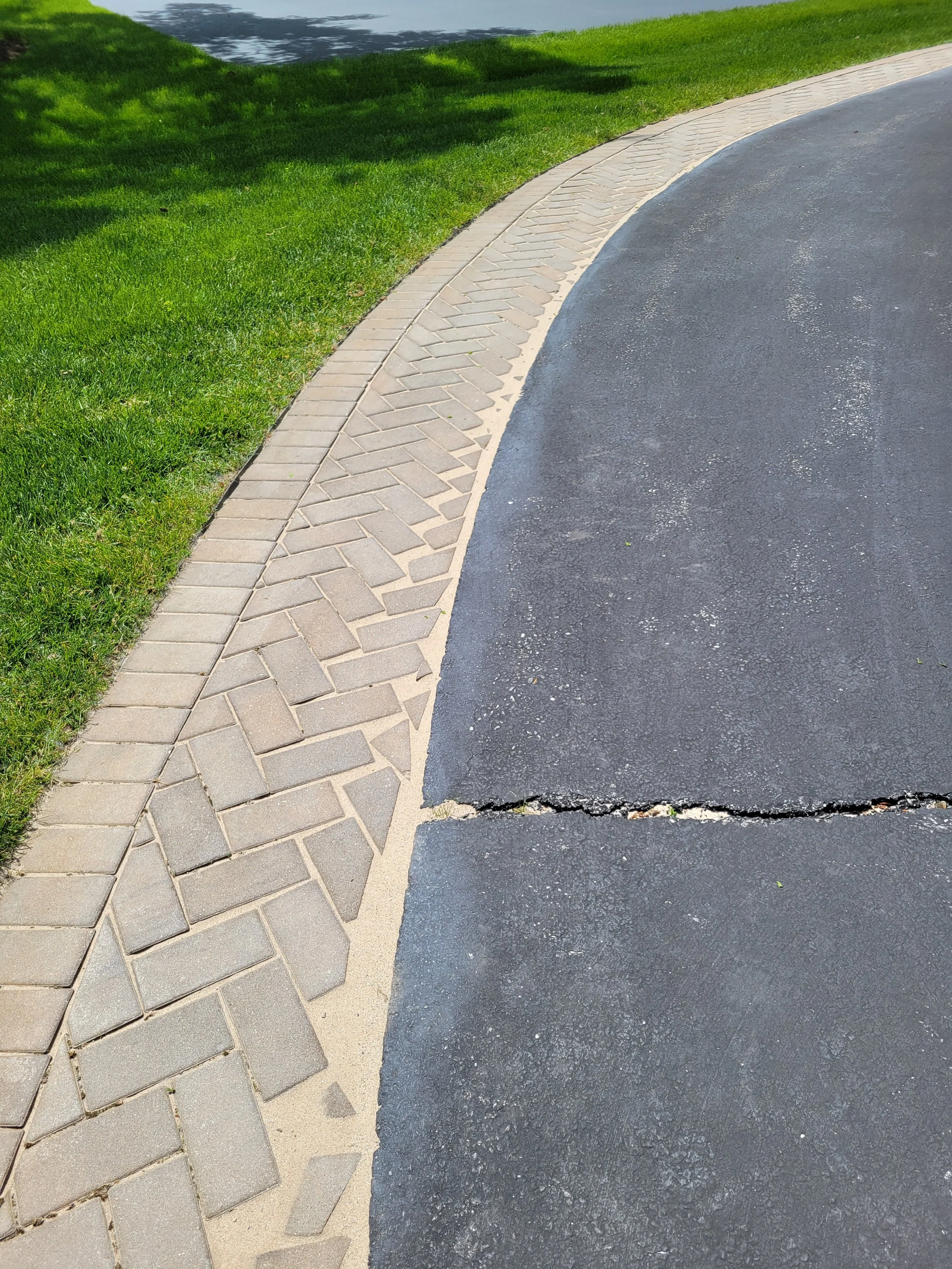 A sidewalk with a brick border running alongside a grassy lawn. A crack is visible in the asphalt surface of the sidewalk.