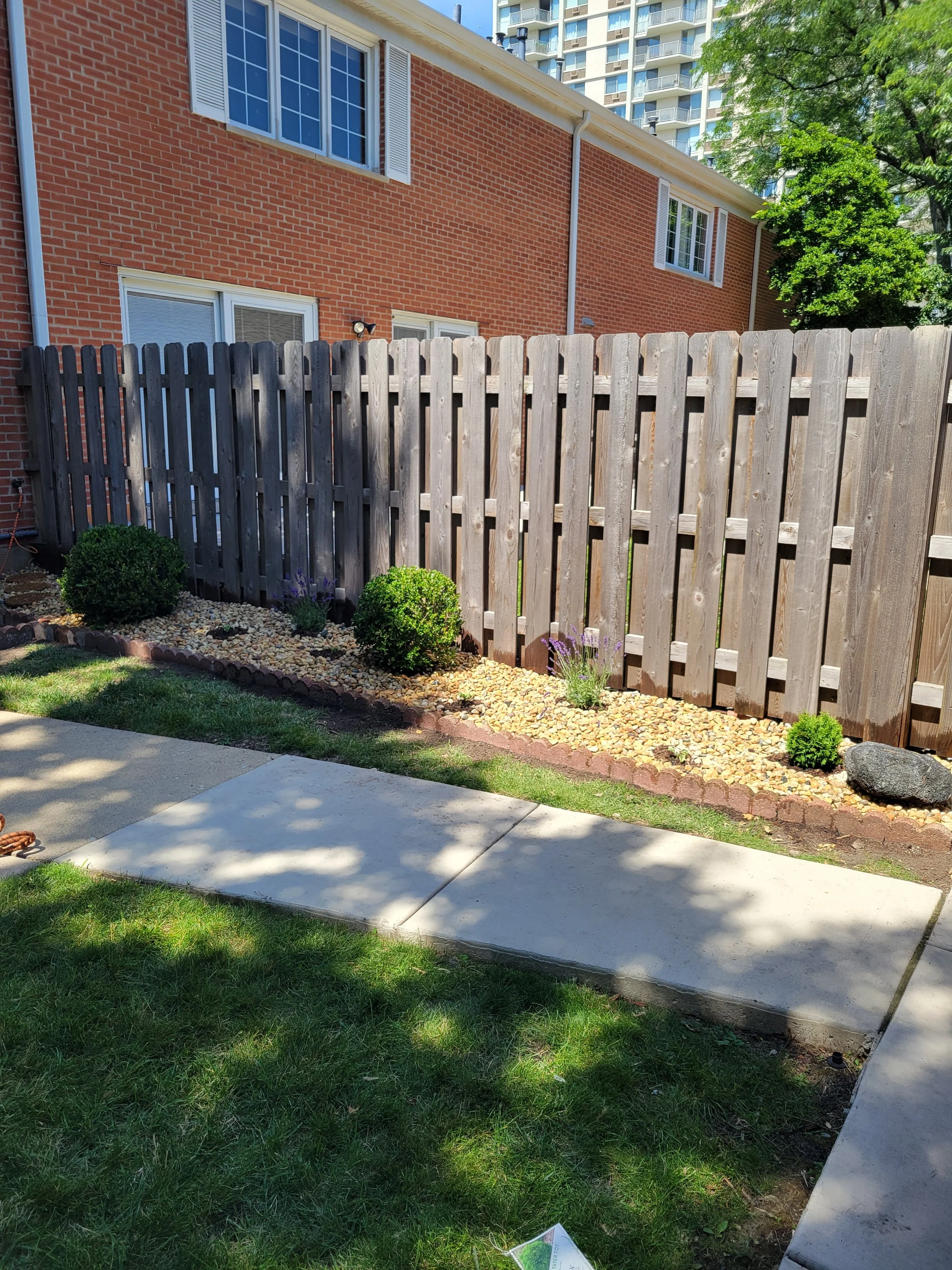 Backyard with a concrete patio, grassy lawn, a wooden fence, small bushes, and purple flowers in a garden bed with decorative rocks.