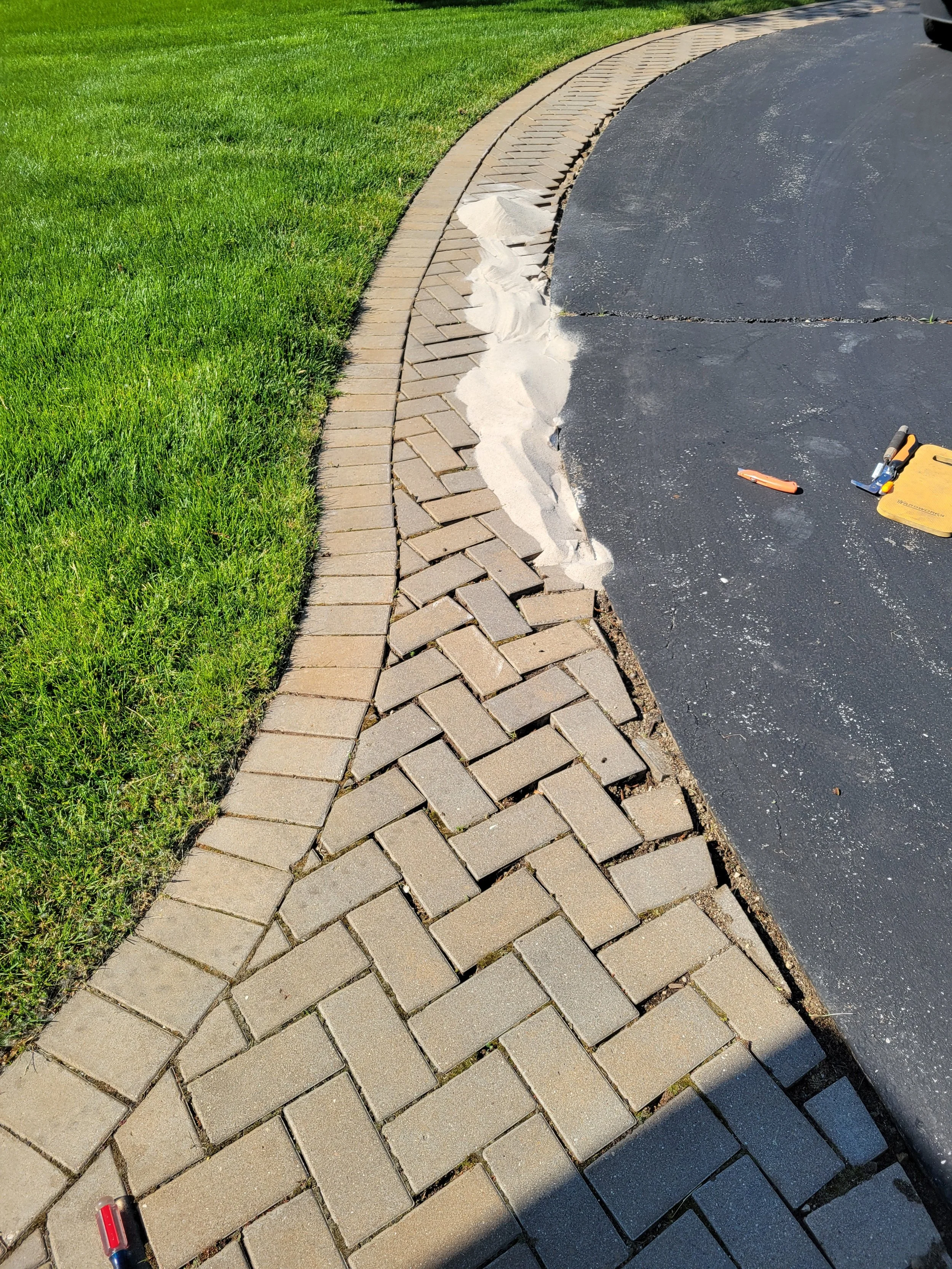 A sidewalk with a new brick border being installed alongside a paved road. There is green grass on one side and construction tools on the other.