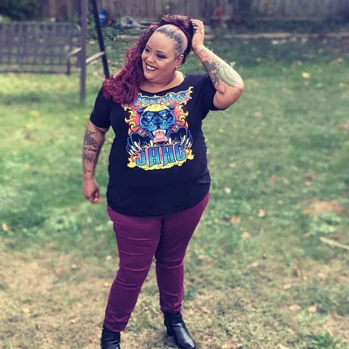 A woman with curly hair and tattoos standing outdoors on grass, smiling, wearing a black t-shirt with a graphic of a tiger and the word 'JAG,' purple pants, and black boots.