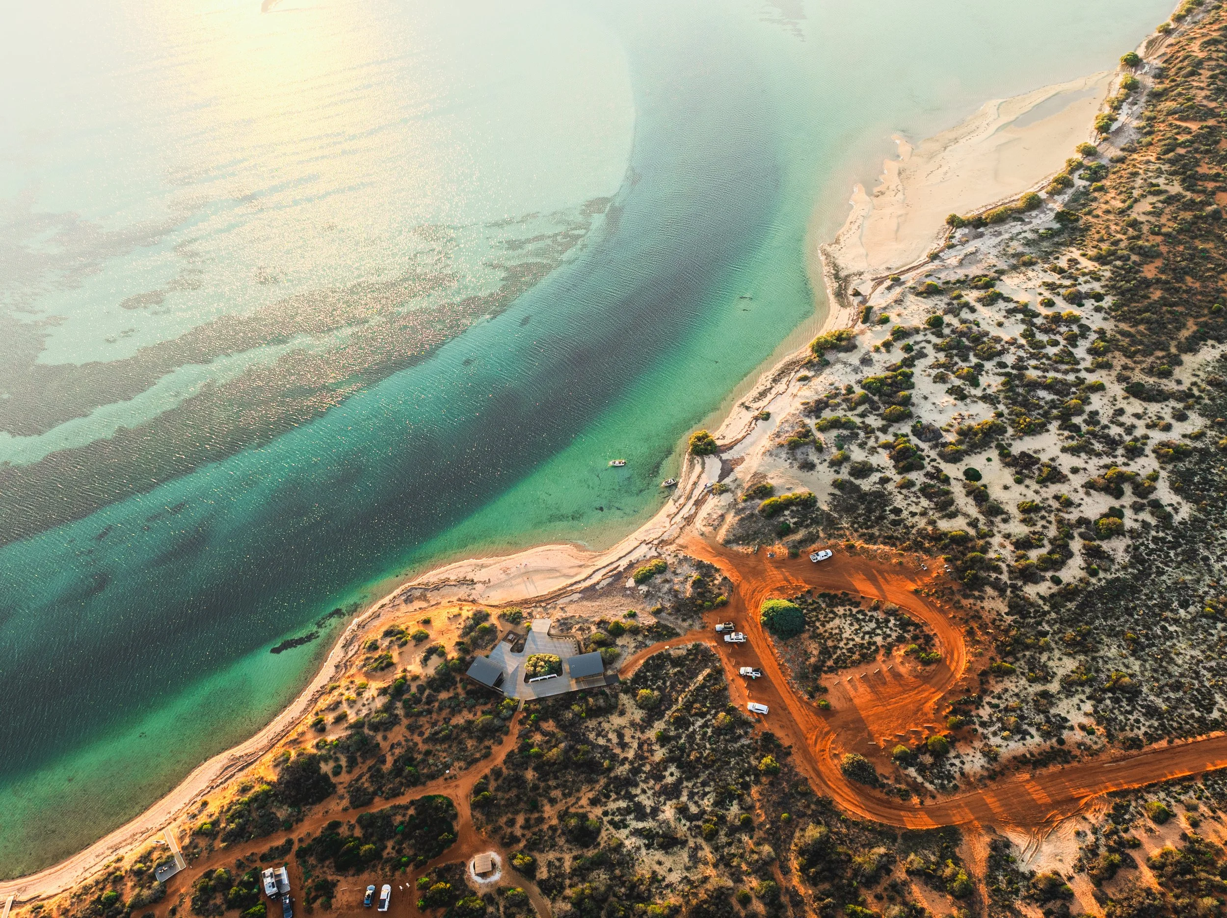 Aerial view of a shoreline with turquoise water and a sandy beach, adjacent to a dry, rocky landscape with scattered bushes and a few structures and vehicles.