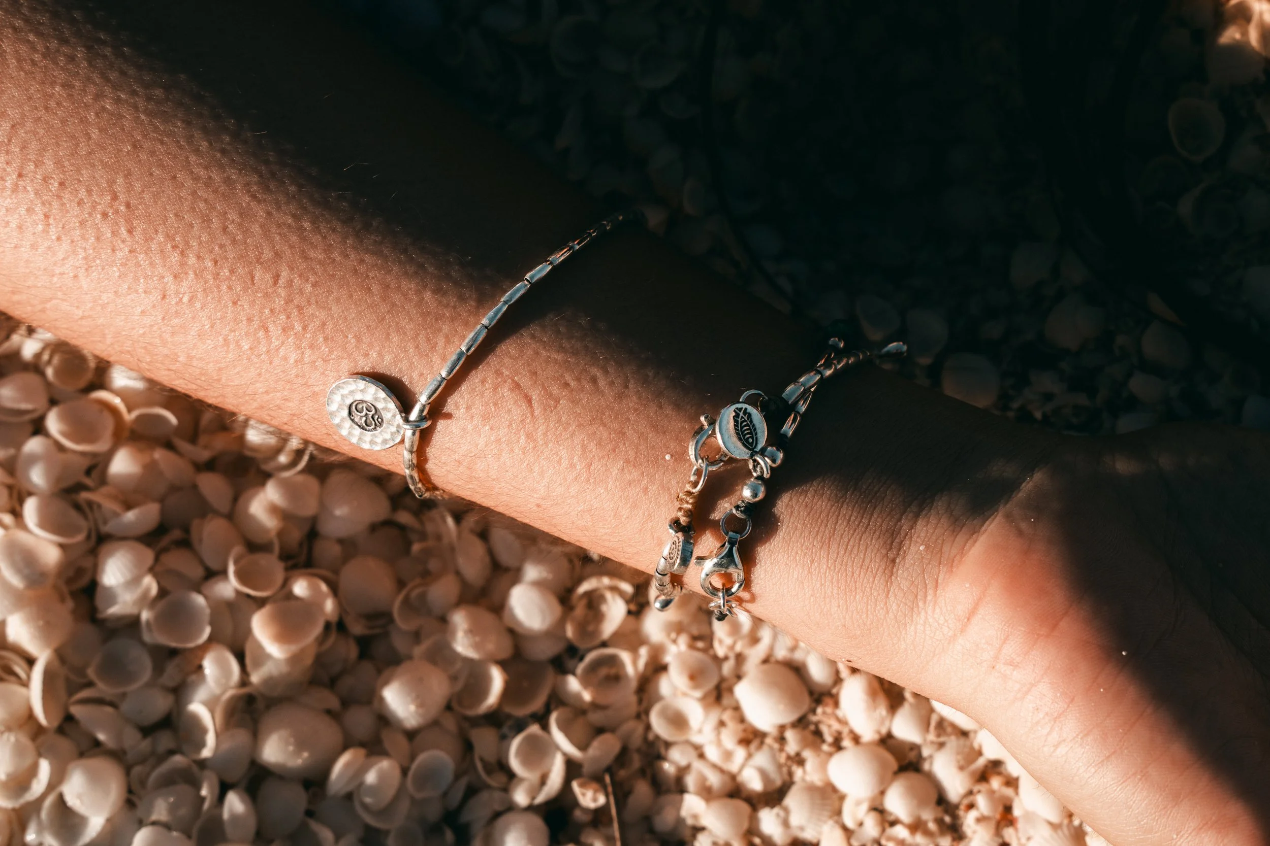 Close-up of a person's arm with two silver bracelets, one with a small round charm, resting on a bed of small seashells.