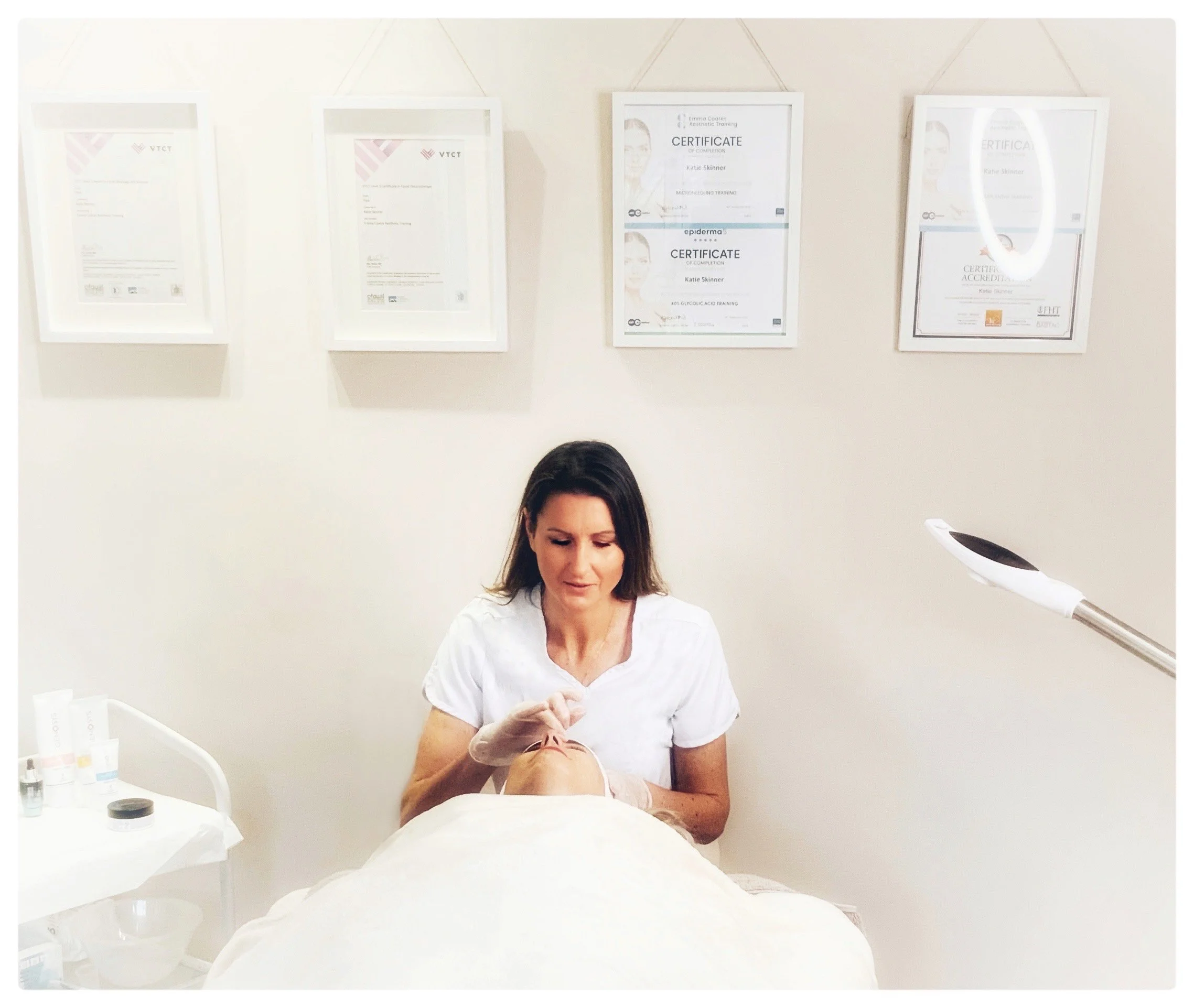 A woman performing a facial treatment on a client lying on a treatment bed in a spa or clinic room decorated with framed certificates on the wall.