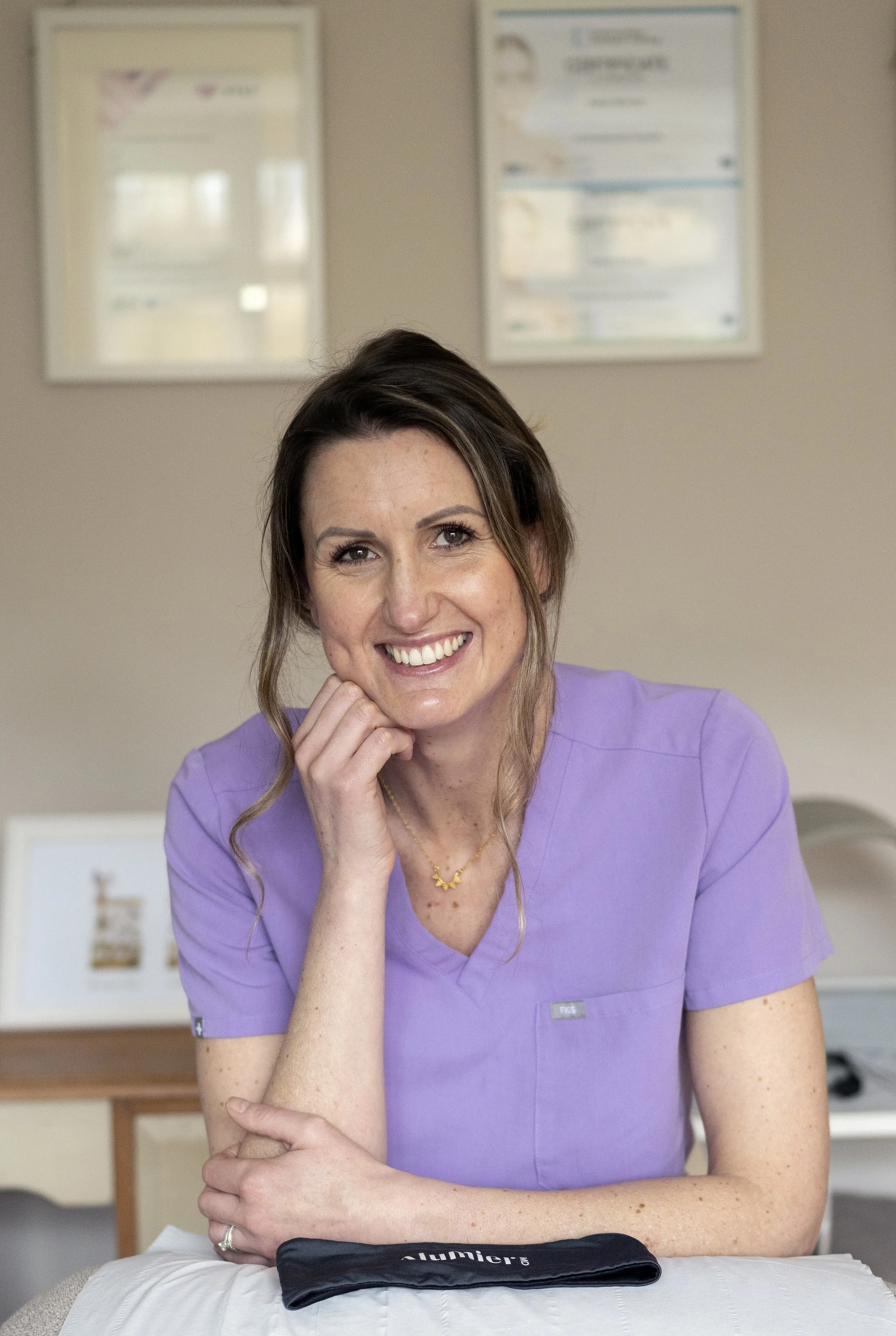 Katie is smiling in purple scrubs sitting at a desk in a skin clinic, with framed certificates on the wall behind her.
