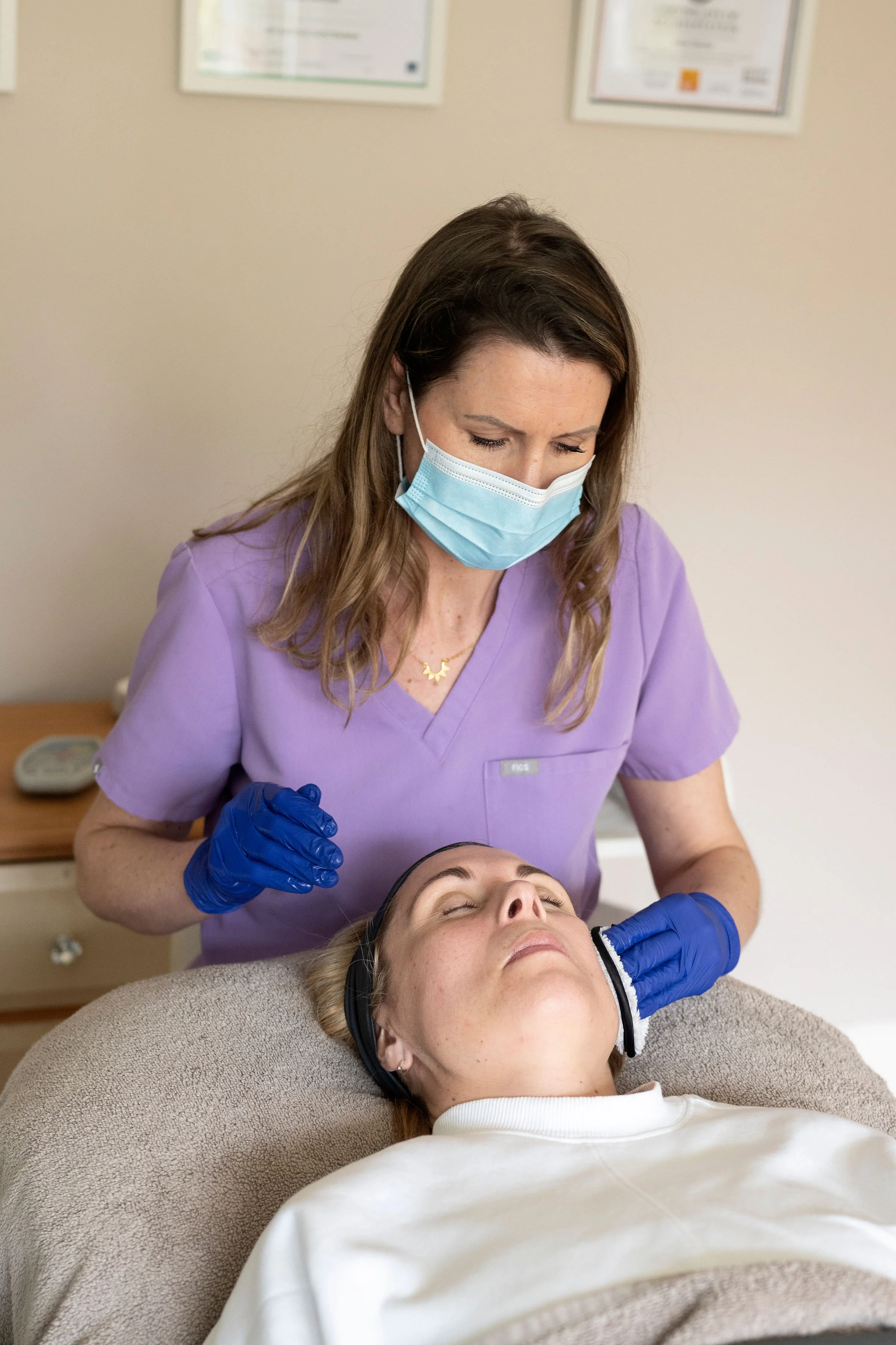 A woman receiving a facial treatment from a healthcare professional in a clinical setting. The professional is wearing purple scrubs, a face mask, and blue gloves, and is gently touching the patient's face as she lies on a treatment bed with her eyes closed.