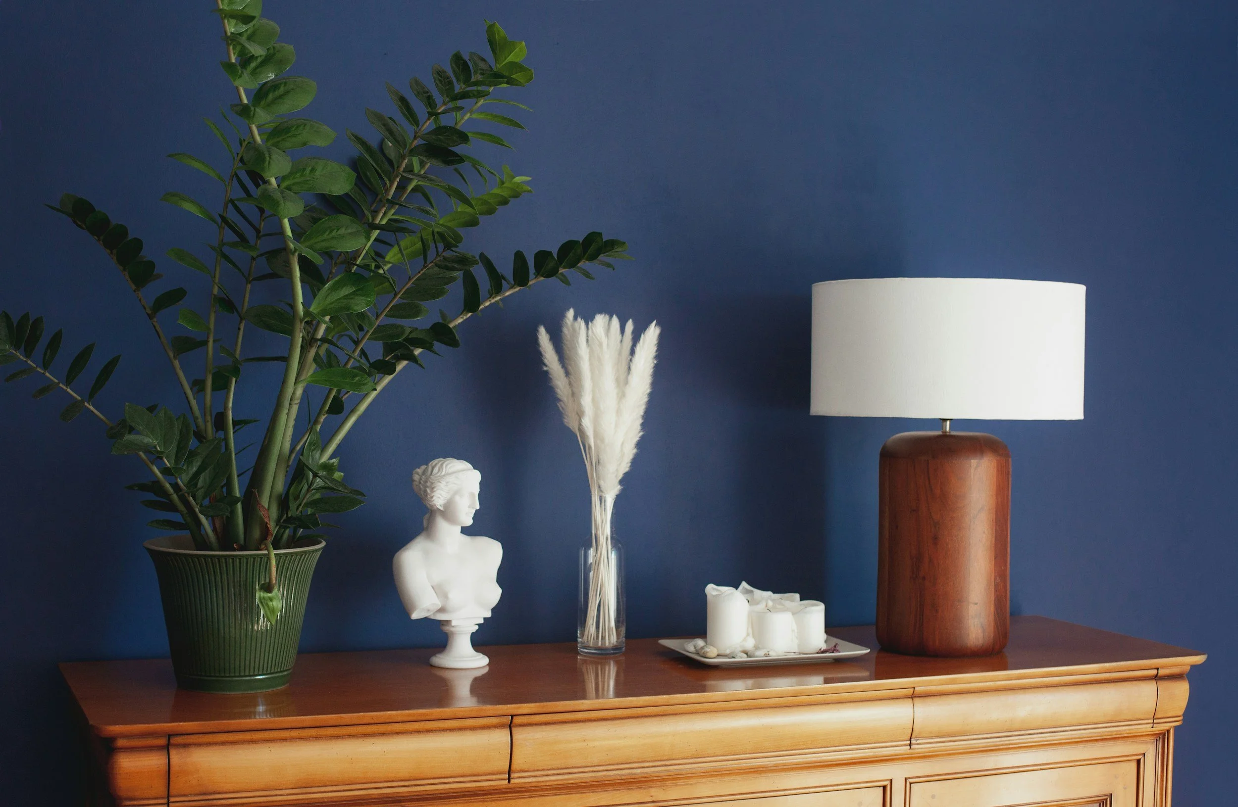 Decorative wooden sideboard against a blue wall, featuring a large potted green plant on the left, a white bust sculpture, a tall glass vase with white feathery plants, a tray of white candles, and a wooden table lamp with a white lampshade.