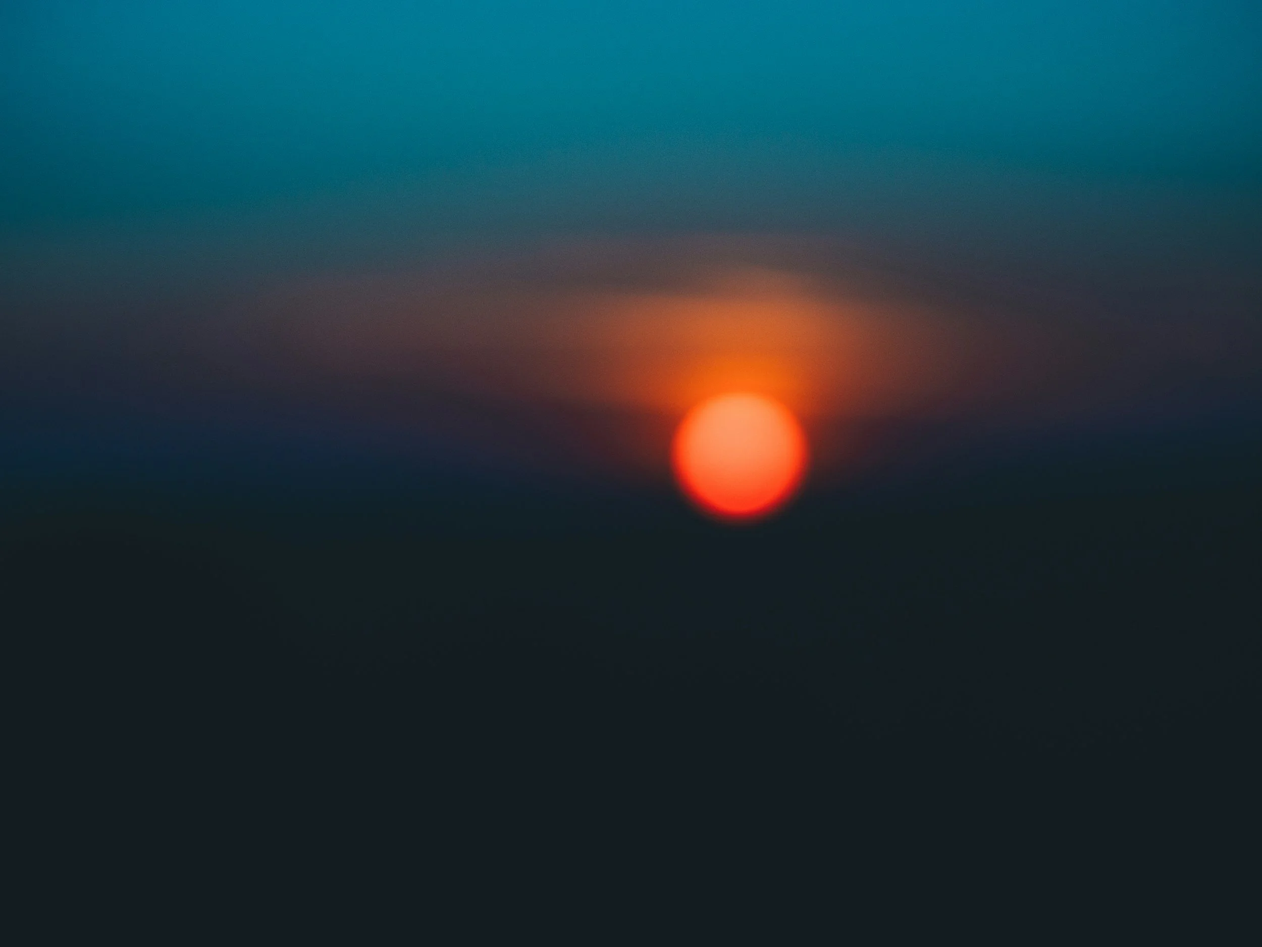 Blurred image of a sunset with a red-orange sun at the horizon and dark landscape below, with a gradient sky above.