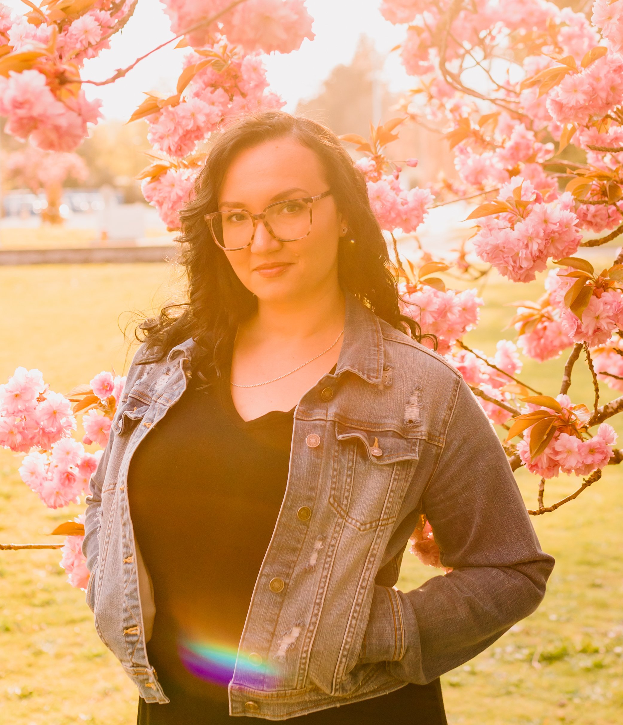 Mila Loskutova, RCC in Vancouver, stands outdoors near pink cherry blossom trees, wearing a denim jacket over a black top, with a slight smile on her face.