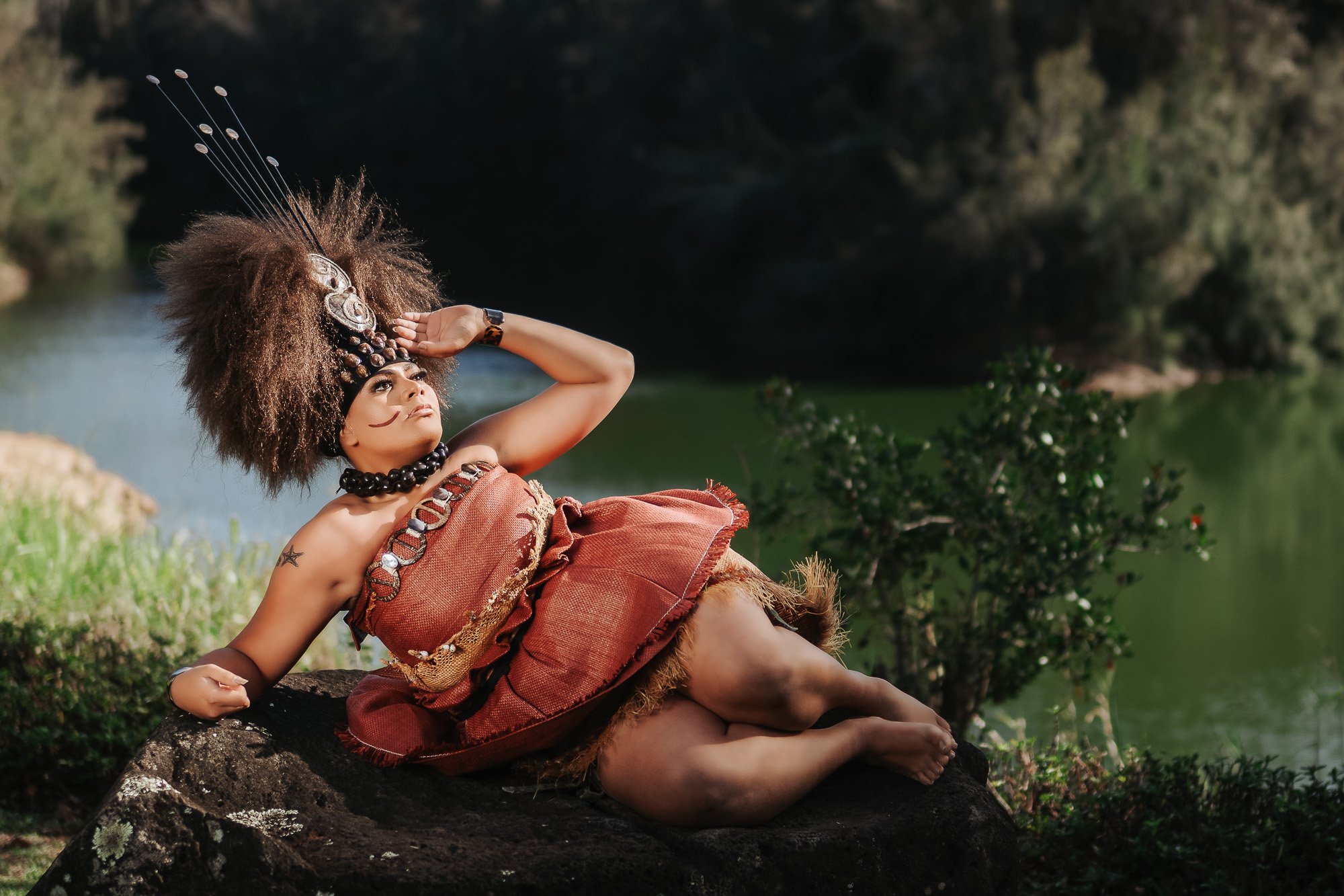 Person dressed in traditional tribal attire, with large feathered headdress, lying on a large rock near a body of water, surrounded by greenery.