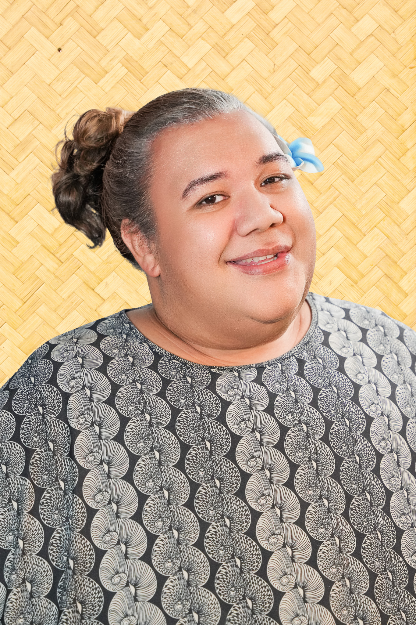 A person with dark hair styled in a bun, wearing a patterned shirt, smiling, with a light blue cloth or bow near the ear, and a woven wooden background.