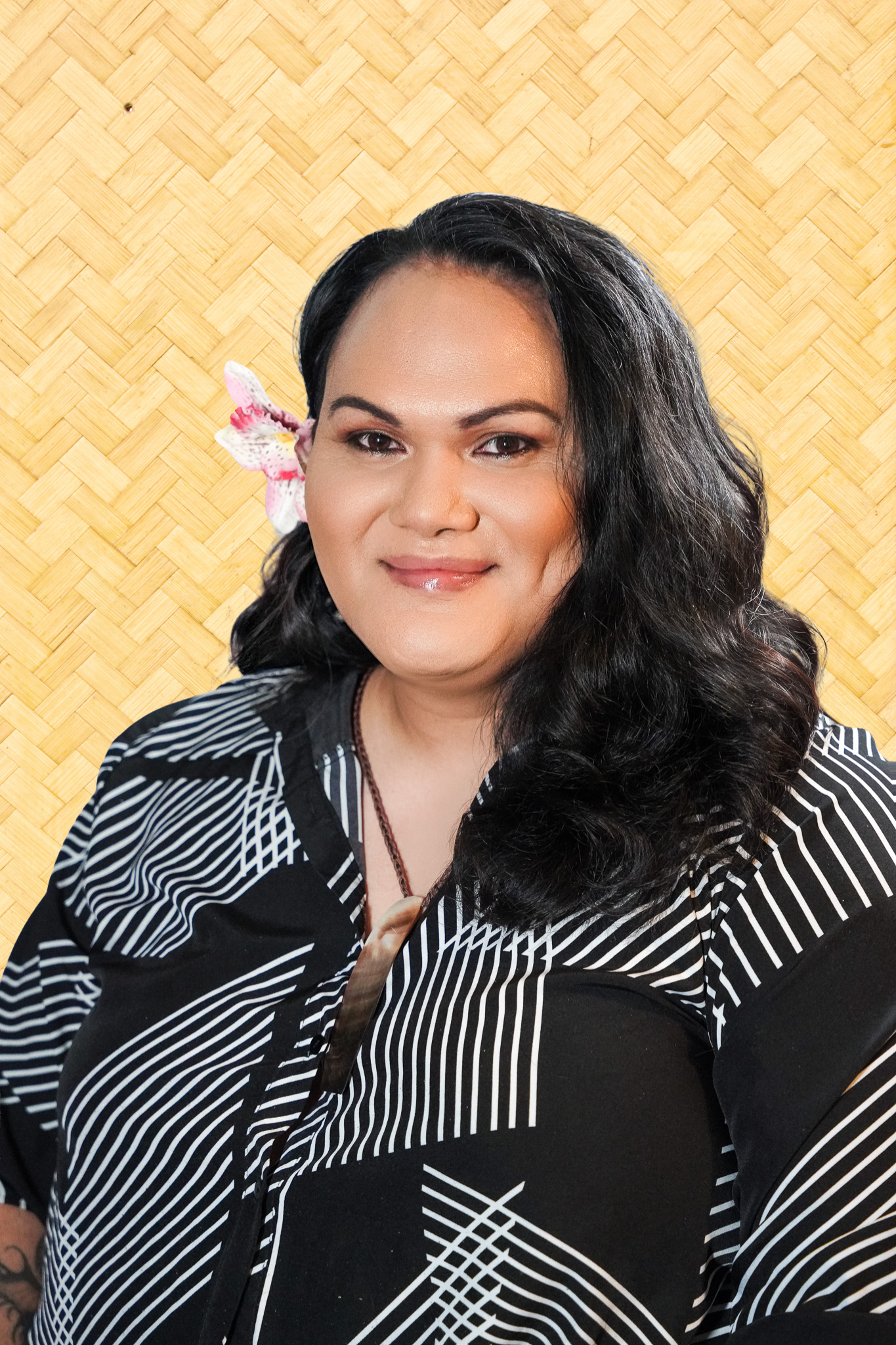 A woman with long dark hair and a flower in her hair, wearing a black and white striped dress, standing in front of a woven bamboo background.