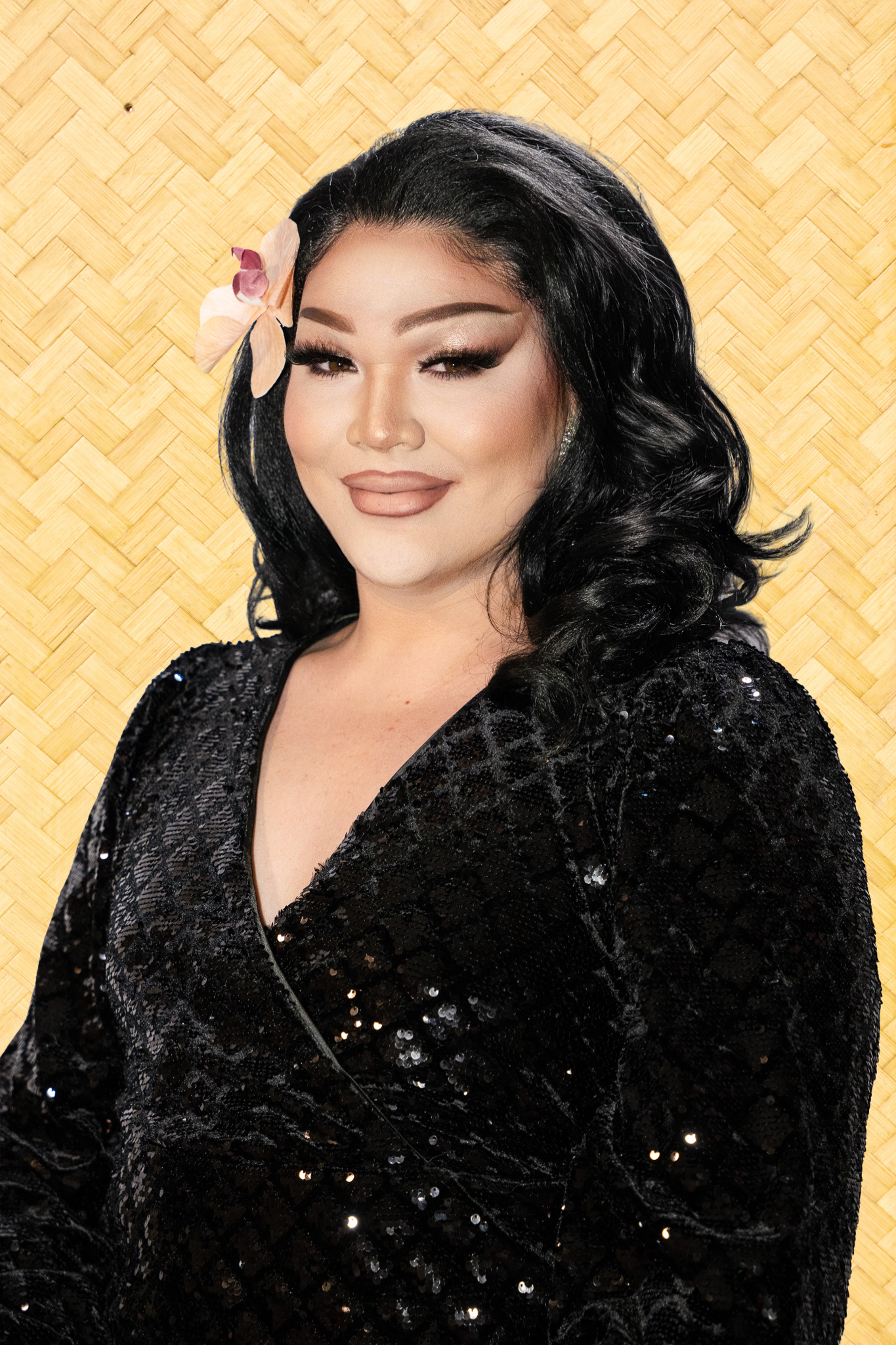 A woman with black hair, makeup with nude lipstick, and a large pink and white flower behind her ear, wearing a black sequin jacket against a woven bamboo background.