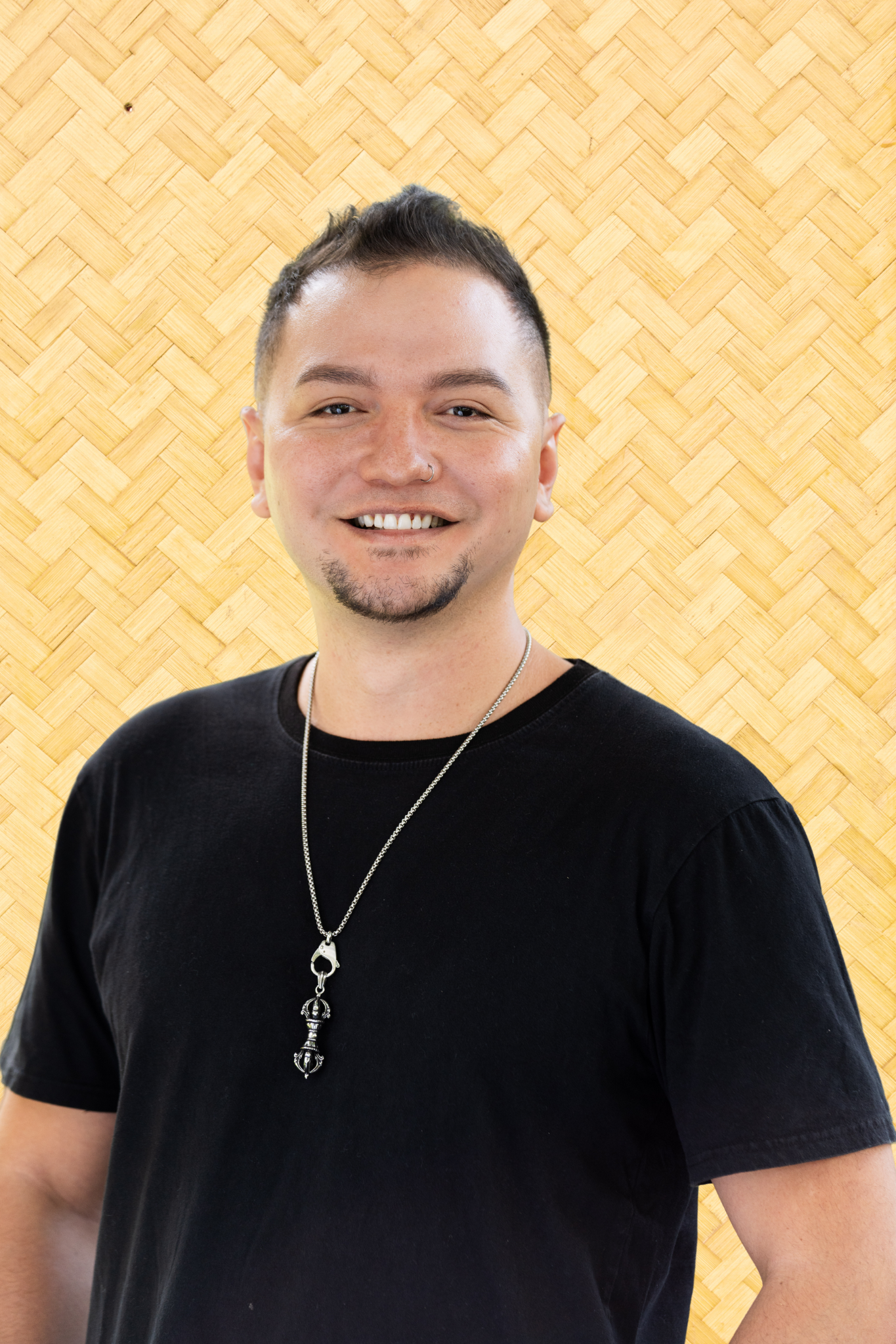 Portrait of a young man with short dark hair, a small goatee, and a nose ring, smiling and wearing a black t-shirt and a silver necklace with a pendant, standing in front of a yellow woven wood background.