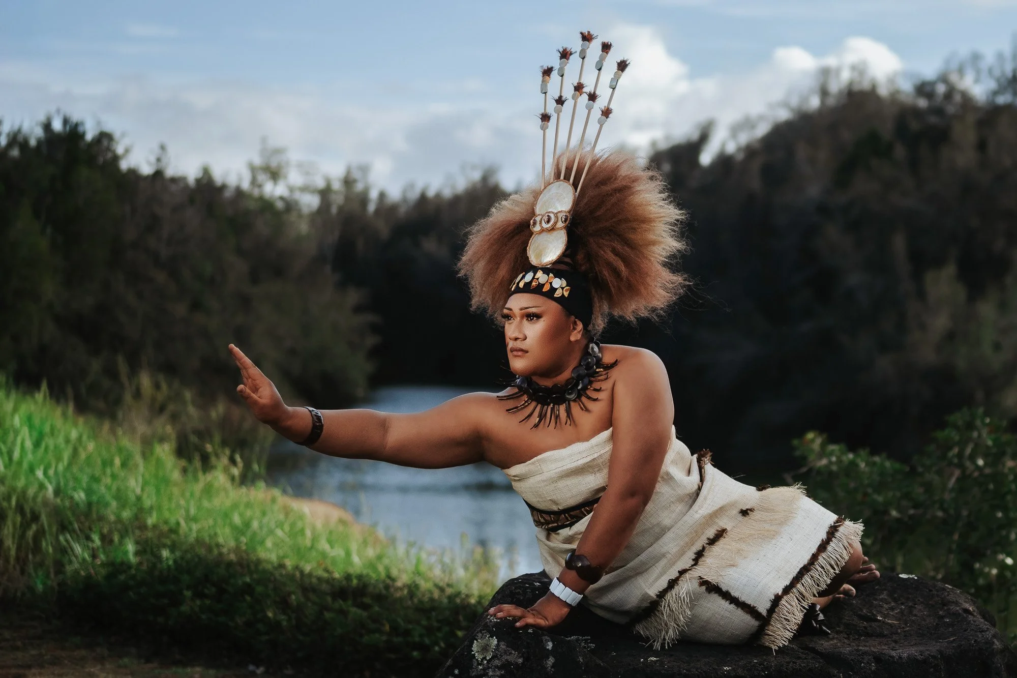 A woman dressed in traditional African attire, with a large feathered headdress, jewelry, and a beige woven cloth, poses outdoors near a river with greenery and rocky cliffs in the background.