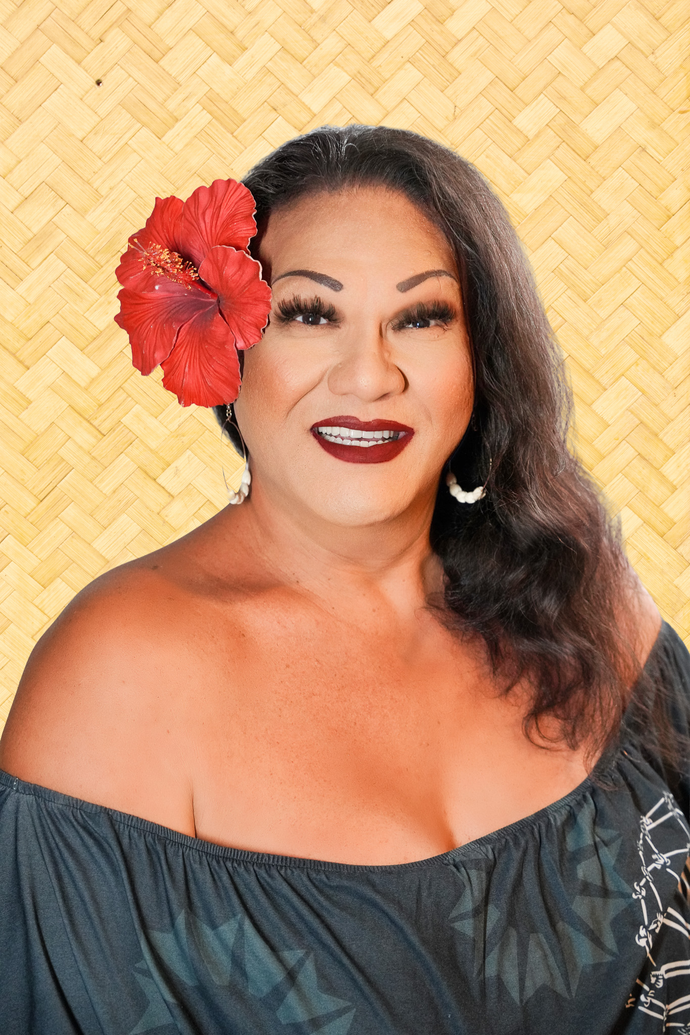 A woman with dark brown hair, wearing makeup including bold red lipstick, and a large red hibiscus flower in her hair. She is smiling and wearing an off-the-shoulder dress with a dark, abstract pattern, standing against a woven background.