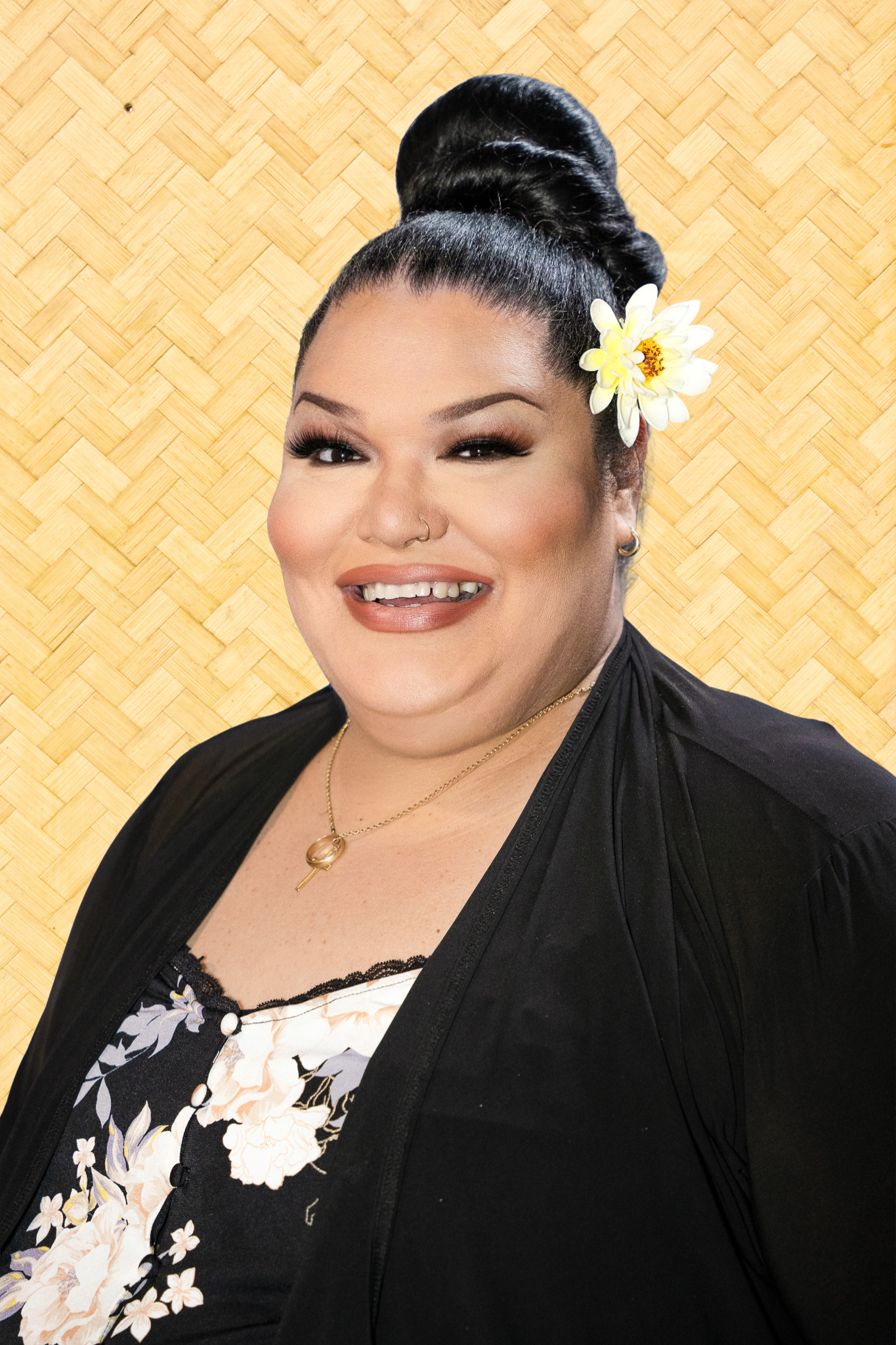A woman with dark hair styled in an elegant updo, with a white flower accessory on the side. She is smiling, showing her teeth, and wearing makeup including eyeliner, eyeshadow, and lipstick. She has a nose ring and small earrings. She is dressed in a black open jacket over a floral top with a black background, white, yellow, and purple flowers. The background is a woven straw pattern.