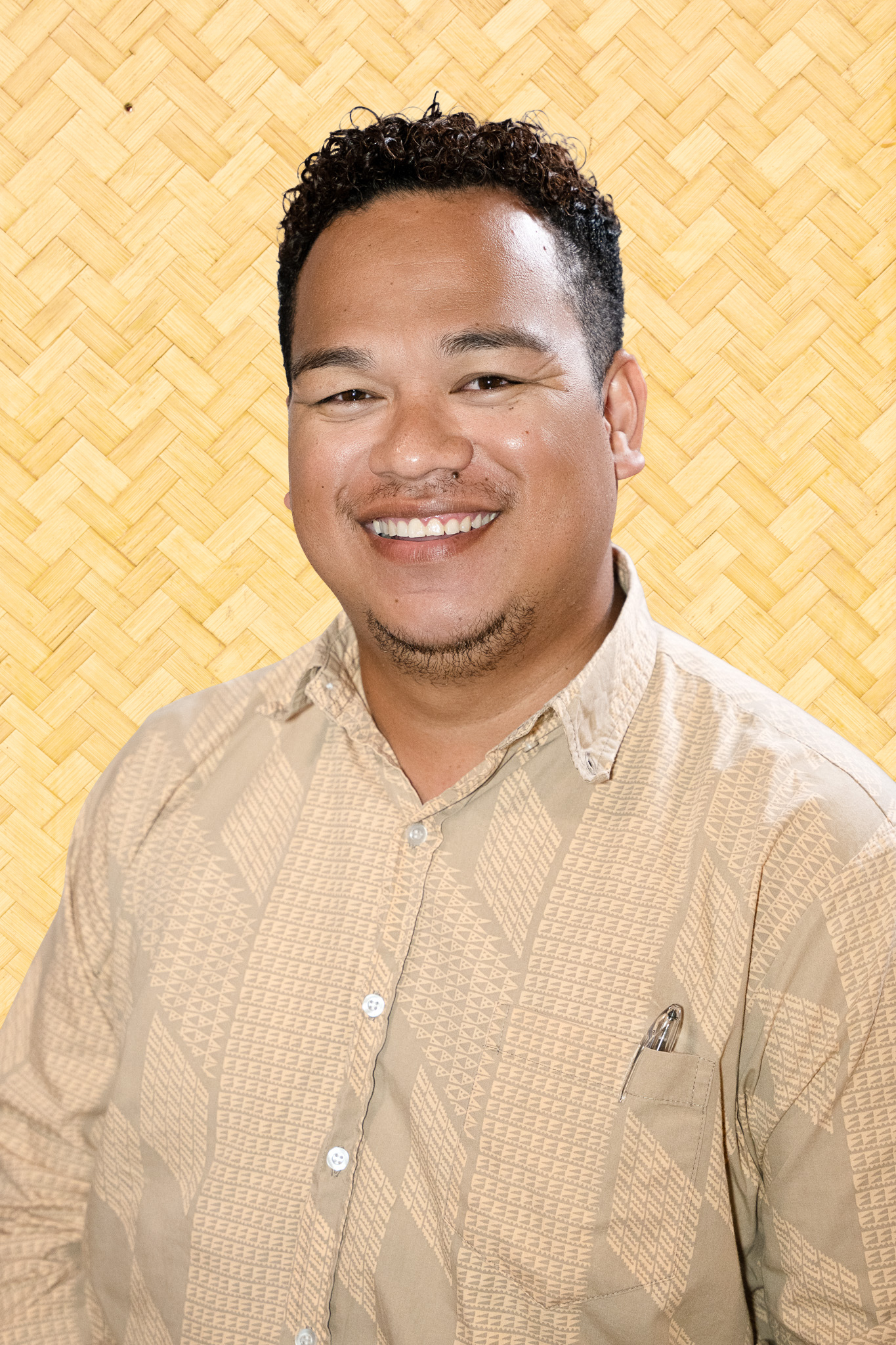 A smiling man with black curly hair and a small goatee, wearing a beige patterned button-up shirt, standing in front of a bamboo-patterned background.