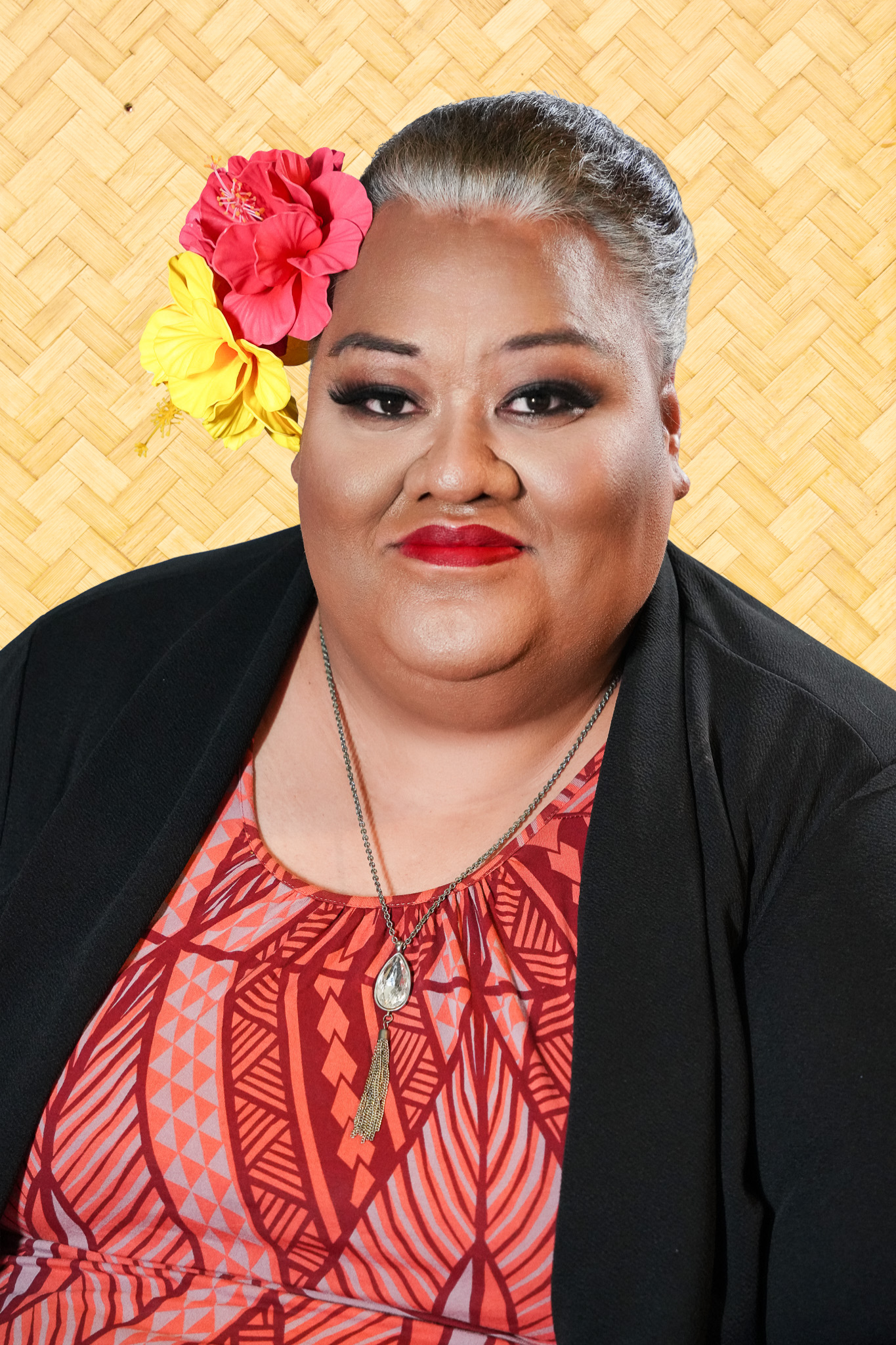 A woman with gray hair styled in a bun, wearing a black blazer and a red patterned dress, with colorful flower accessories in her hair, standing against a bamboo textured background.