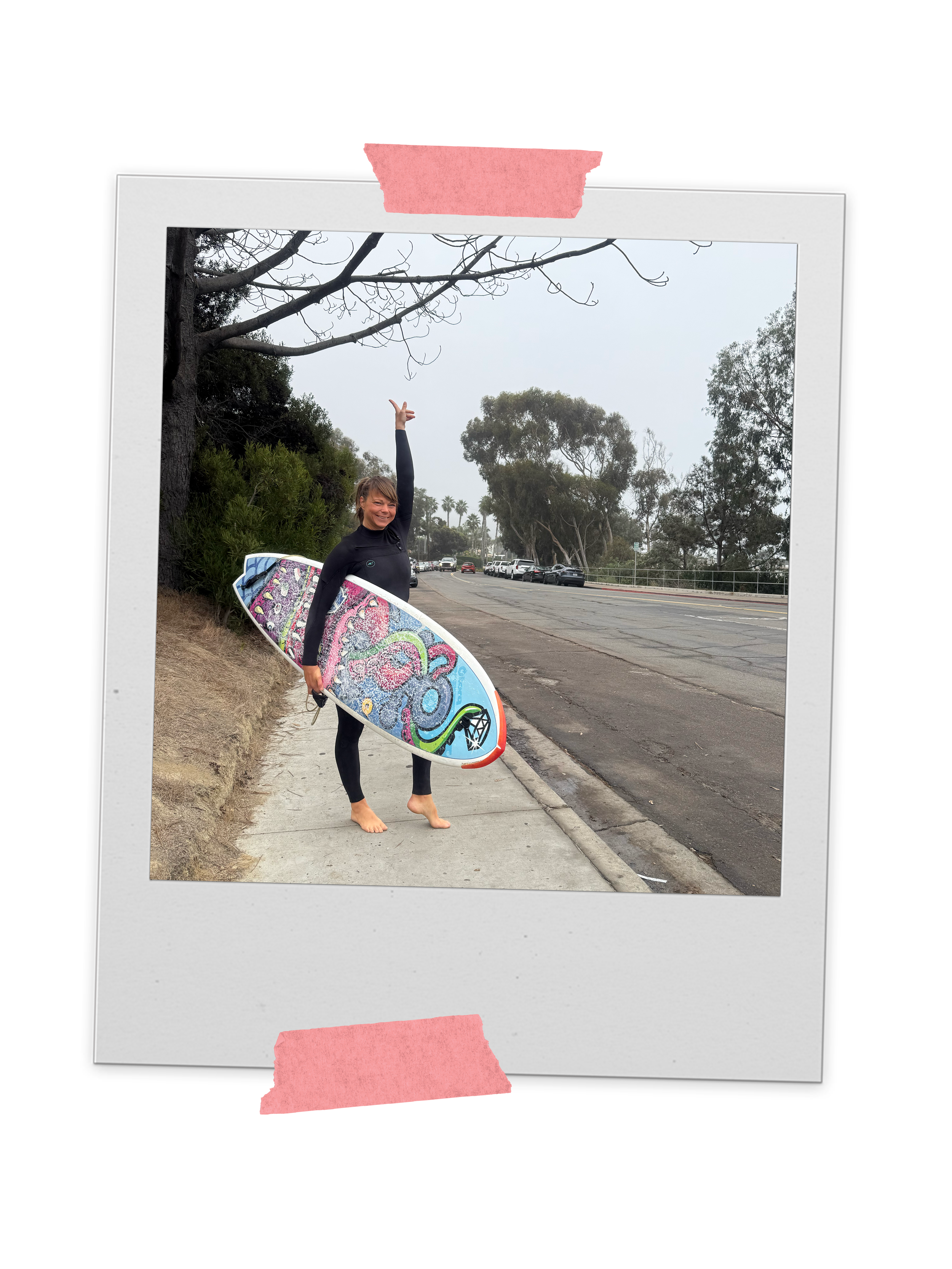 Woman in black wetsuit holding a colorful surfboard on sidewalk near trees and parked cars on an overcast day.