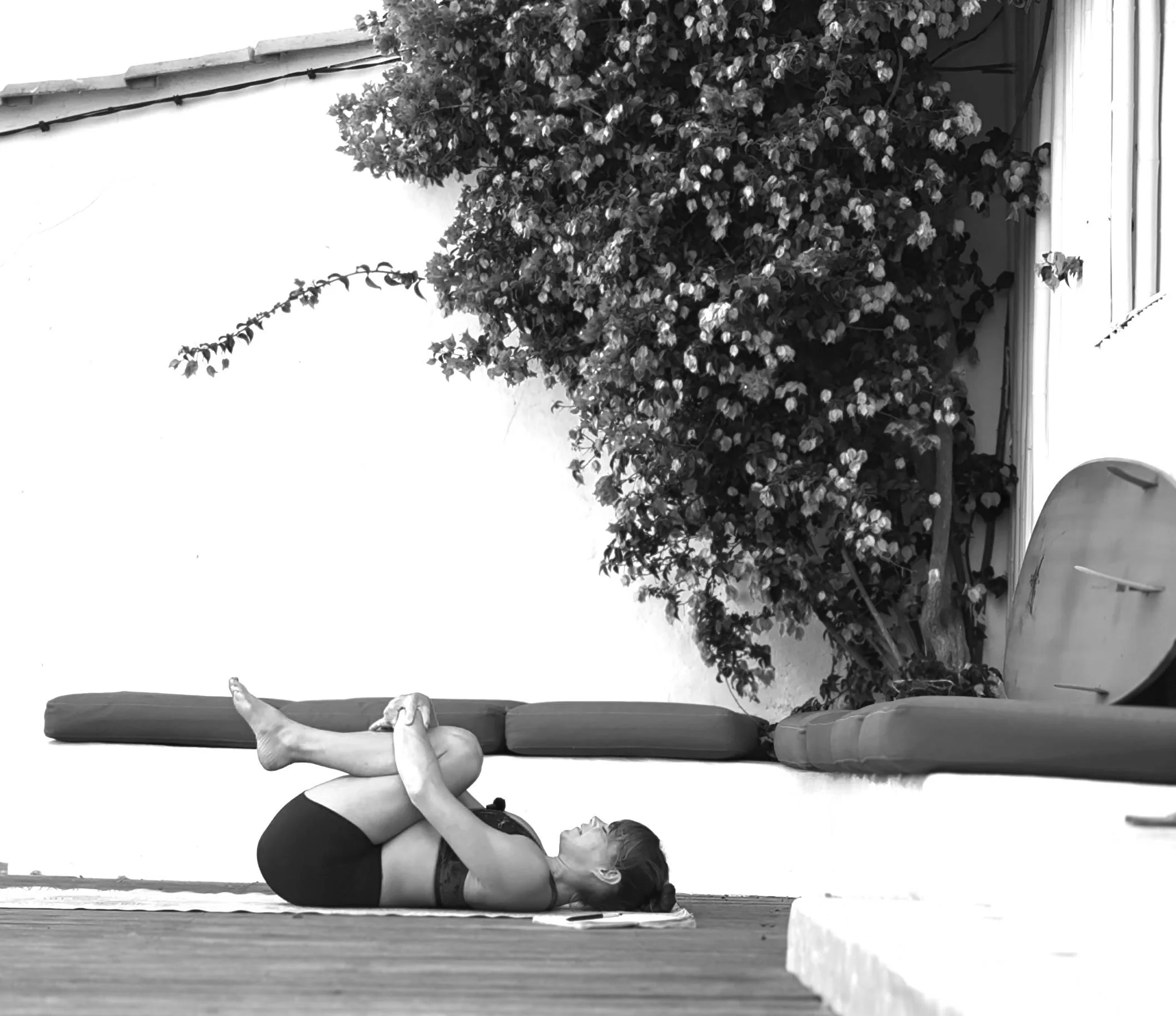 A woman practicing yoga indoors lying on her back with knees bent and hugging her legs, with a large flowering bush and a folded surfboard nearby.