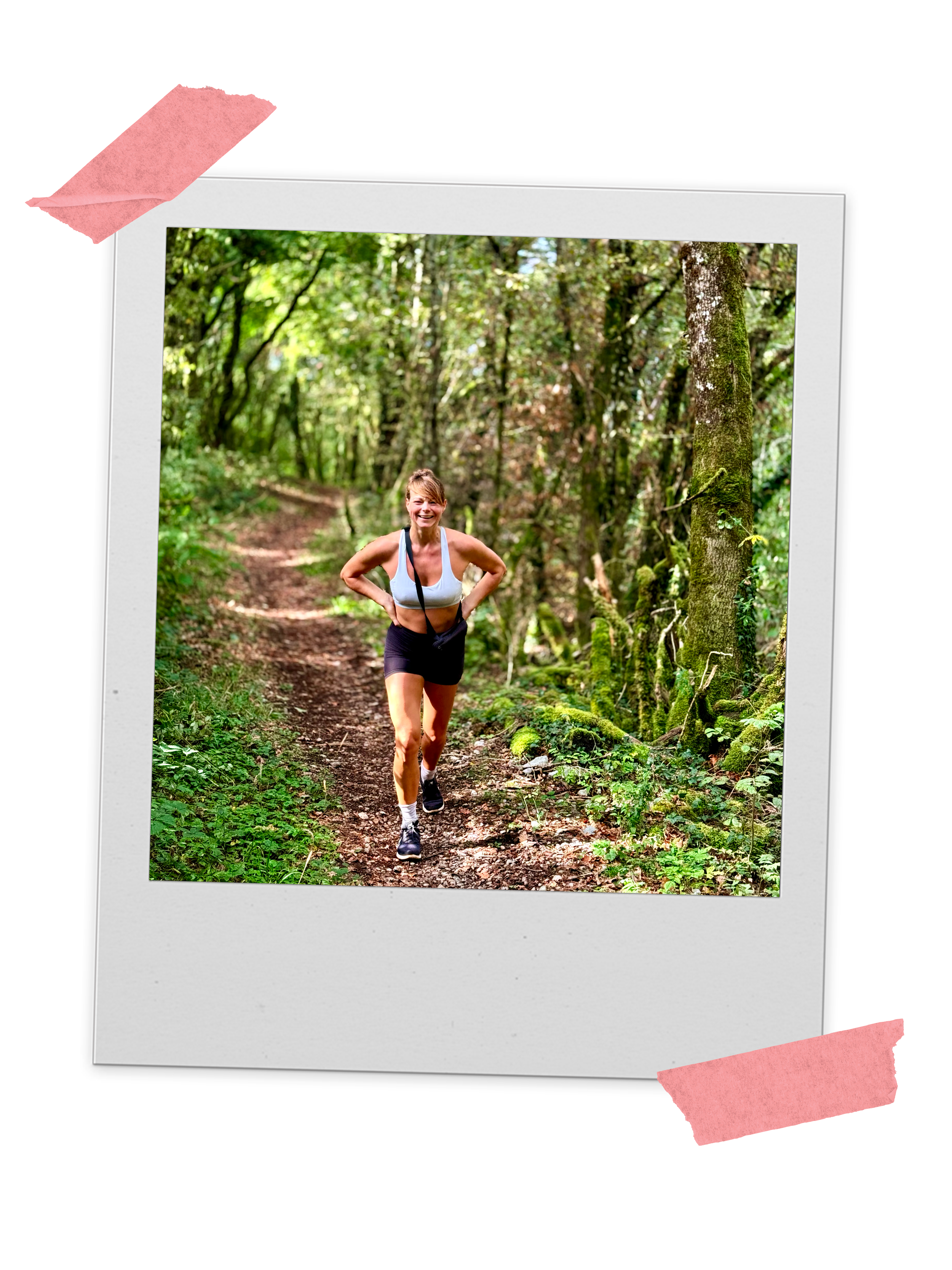 A woman running on a trail through a wooded forest, smiling, wearing a white tank top, black shorts, and running shoes.