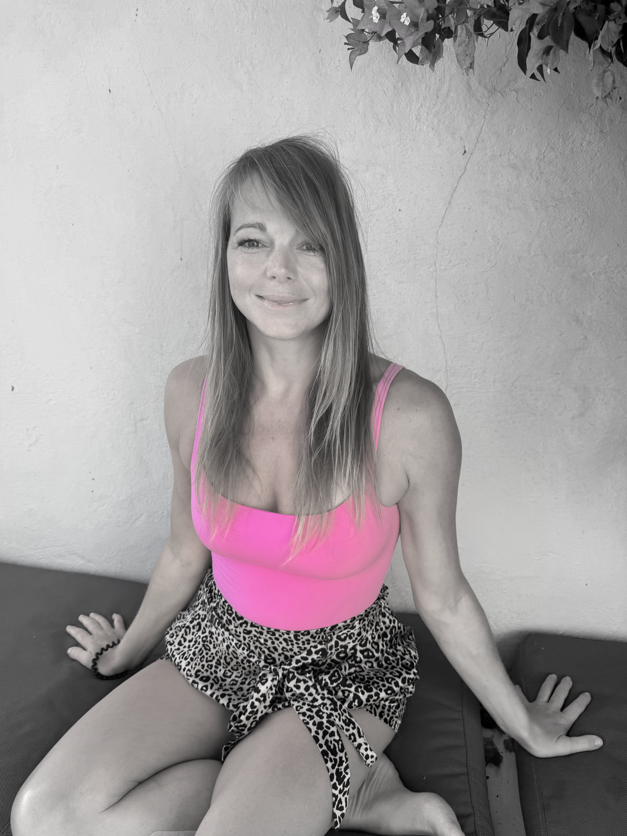 A woman with long hair, smiling, wearing a pink tank top and leopard print shorts, sitting on a cushioned surface against a white wall with some greenery at the top right corner.