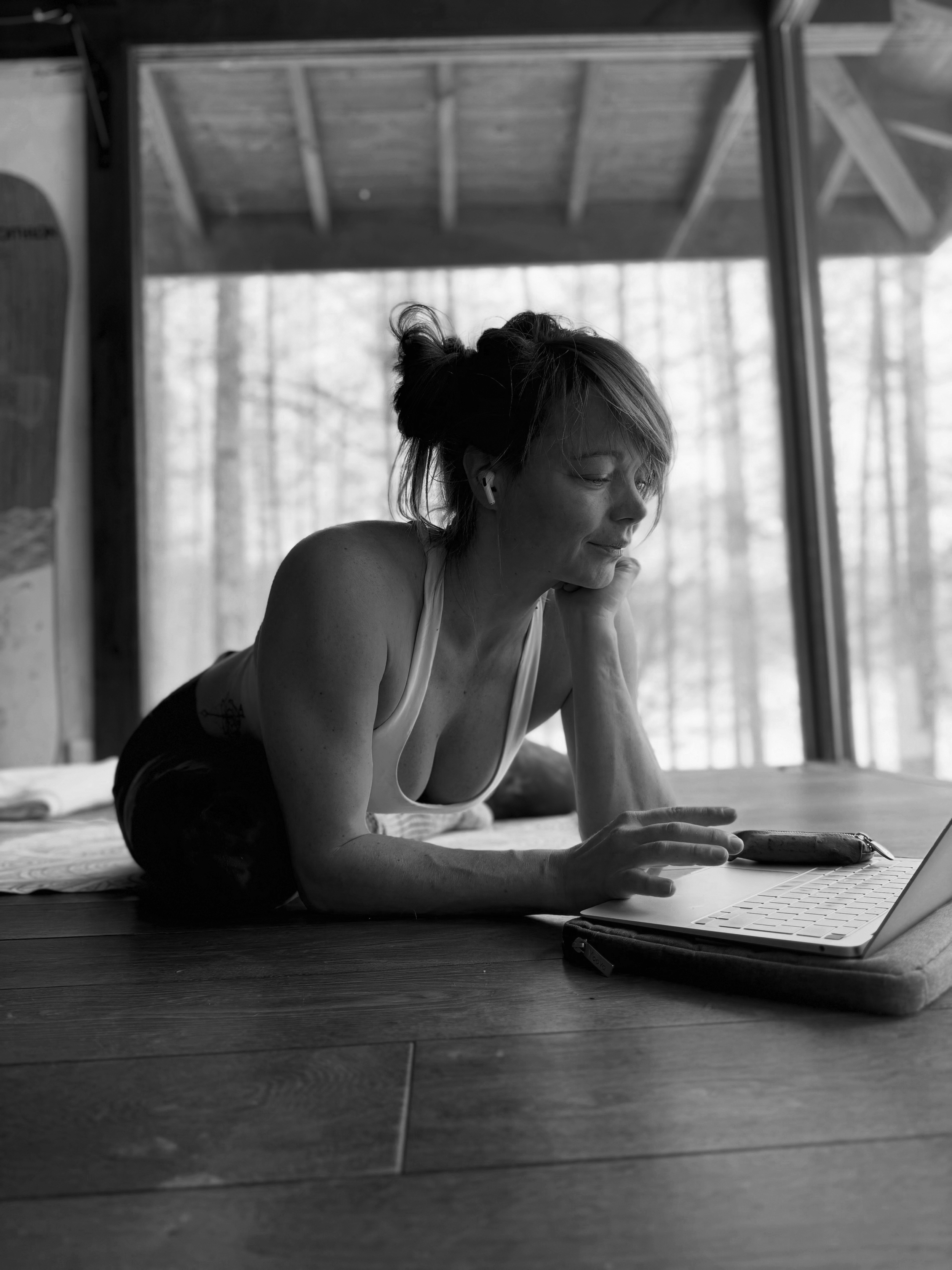 A woman with short hair in workout clothes lying on her stomach on the floor, using a laptop and wearing wireless earbuds, with a wooded window backdrop.