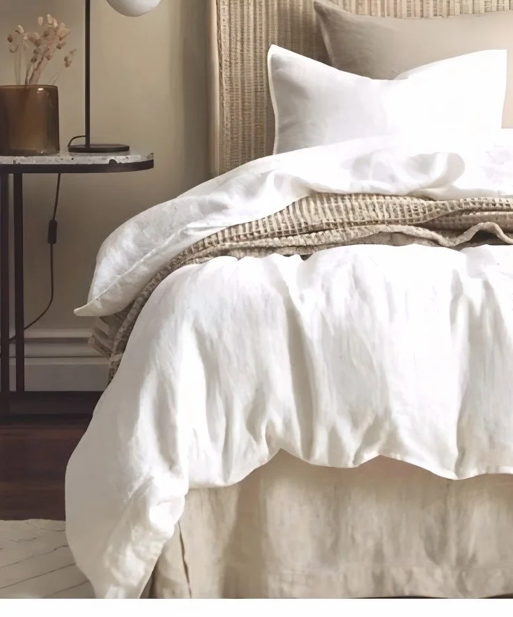 A neatly made bed with white and beige bedding next to a bedside table with a lamp and decorative vase.