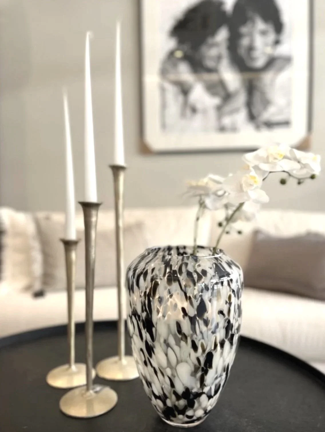 Decorative glass vase with black and white abstract pattern, holding white orchids, placed on a black tray with three silver candlesticks in the background, and a blurred portrait photograph on the wall.