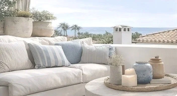 Outdoor patio with white sofa and decorative pillows, round coffee table with candles and vases, in front of a view of palm trees, rooftops, and the ocean.