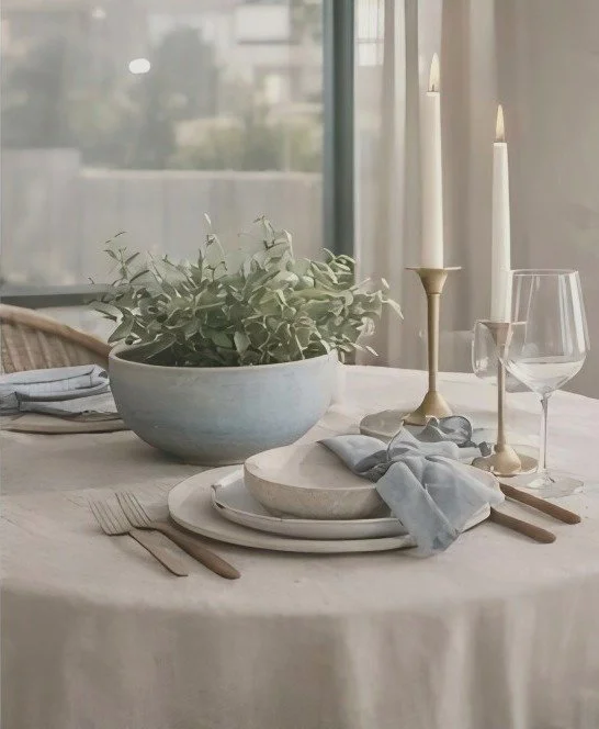 Elegant dining table setup with white tablecloth, a large bowl of green foliage, white plates, gray napkins, cutlery, a wine glass, and tall candle holders with lit candles near a window.