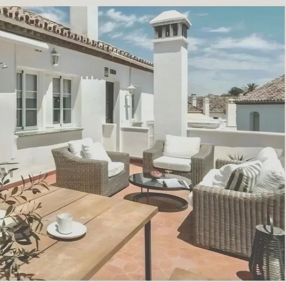 Outdoor patio with white walls, wicker chairs with cushions, a black coffee table, and a wooden table with a cup and saucer, under a partly cloudy sky.