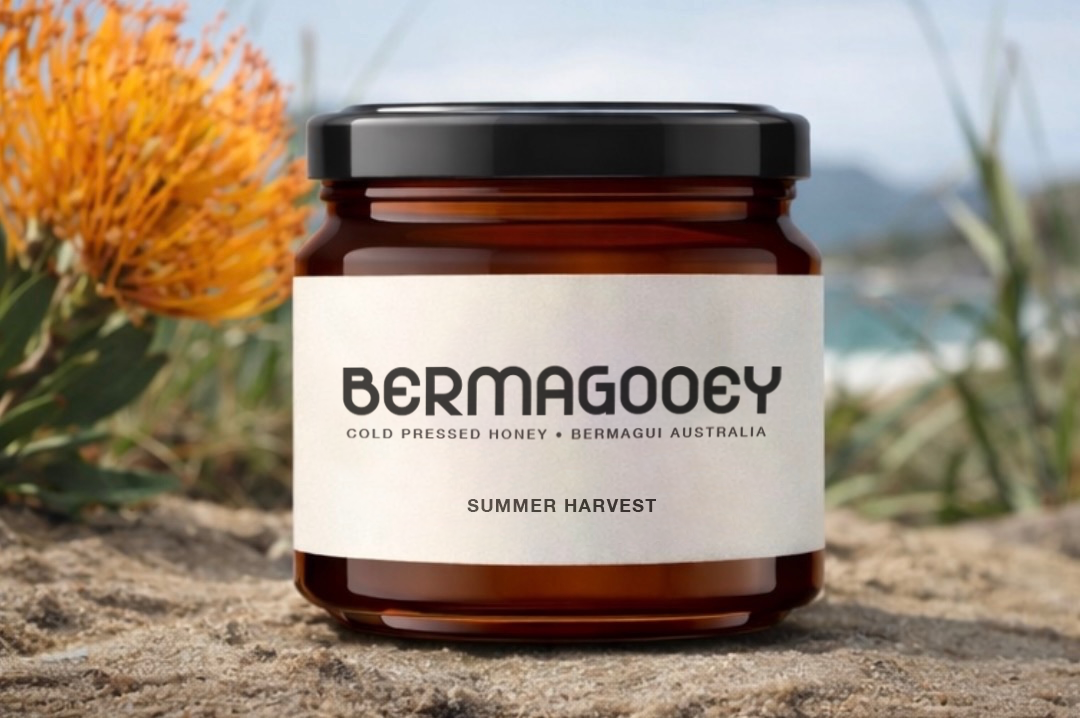 A jar of Bermagooey honey labeled "Summer Harvest" placed on a rocky surface outdoors with orange flowers and blurred scenery in the background.