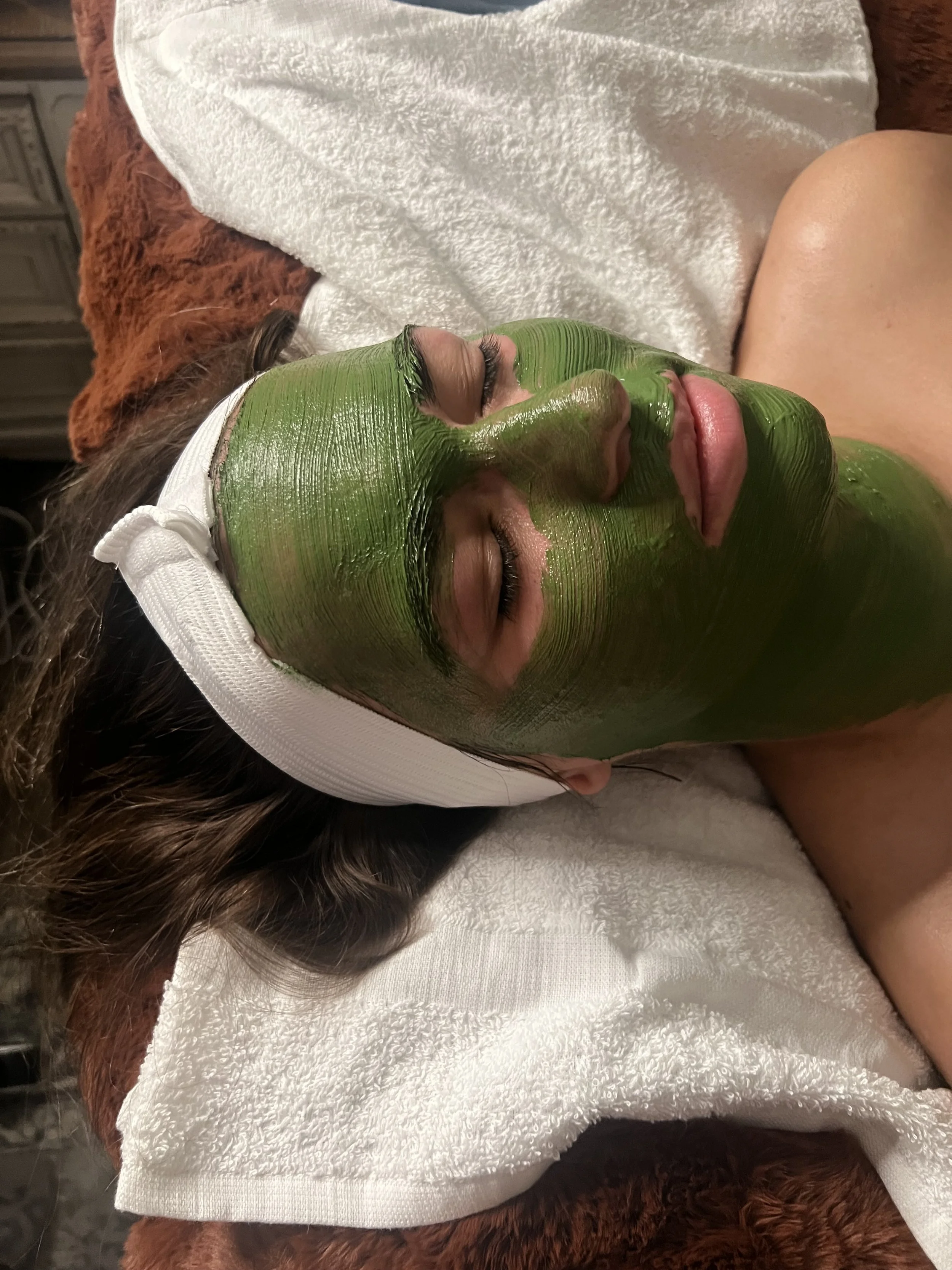 A woman with a green facial mask, lying on a towel with her eyes closed, wearing a white headband, in a spa or skincare setting.