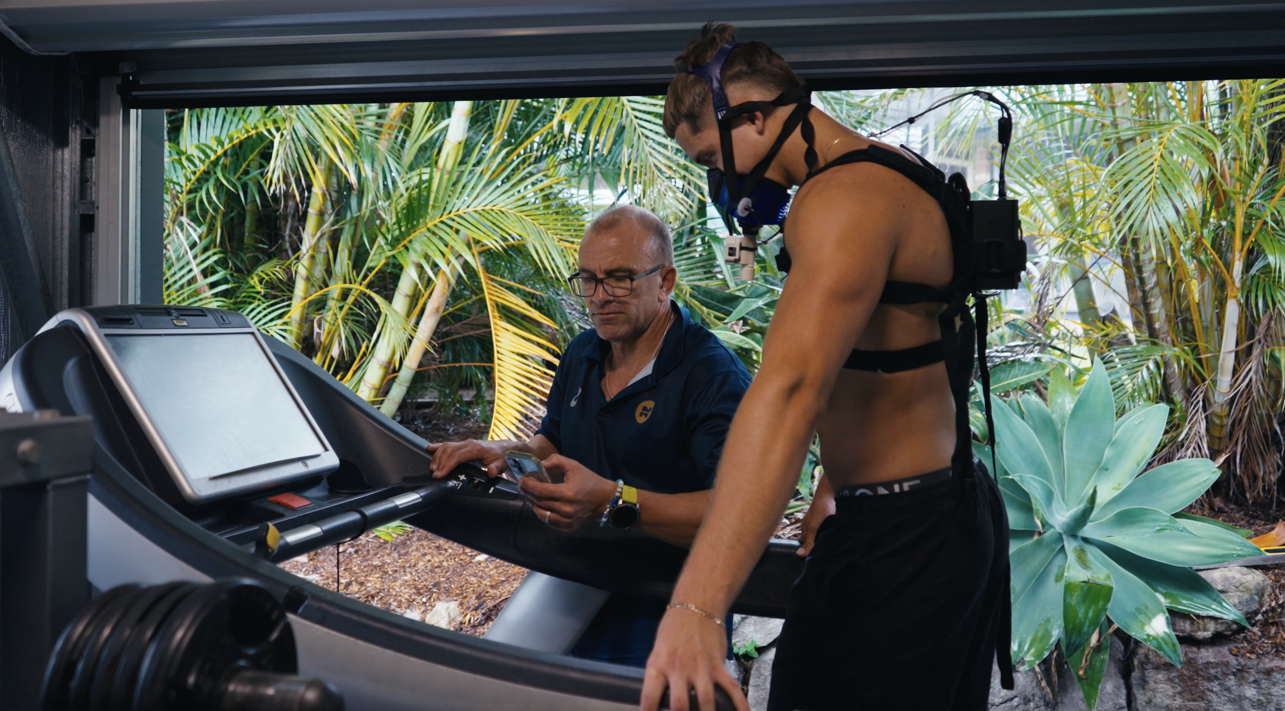 Two men, one older wearing glasses and a navy shirt, and a younger man in athletic gear, are on a treadmill with a tropical garden view in the background.