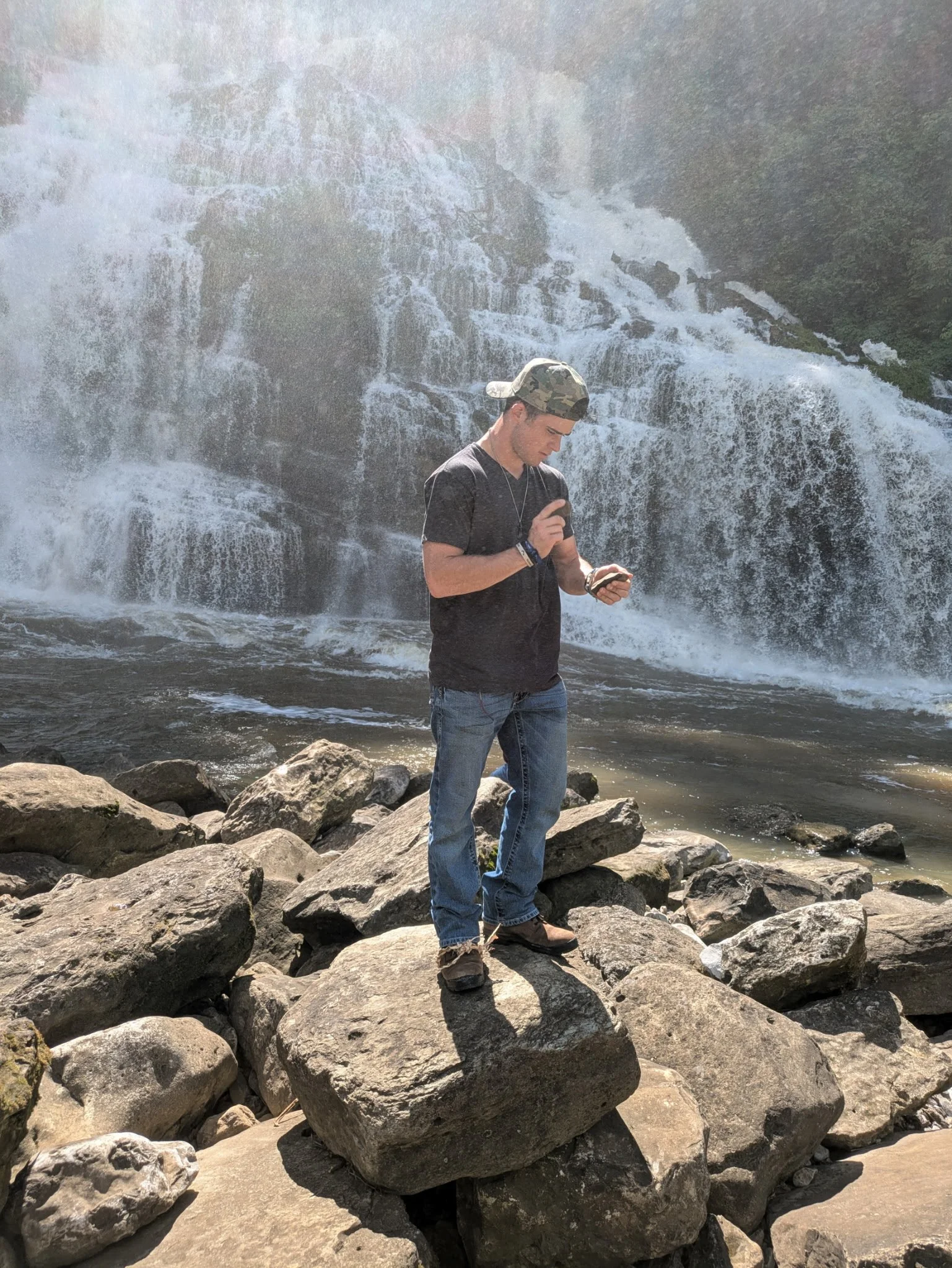 A young man in a black t-shirt and jeans standing on rocks near a waterfall, knapping a rock.