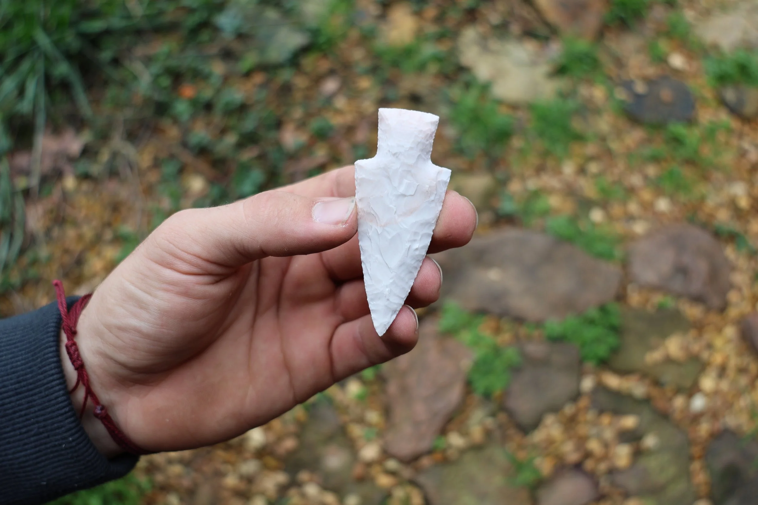 Person holding a white flint arrowhead outdoors amid rocks and greenery.