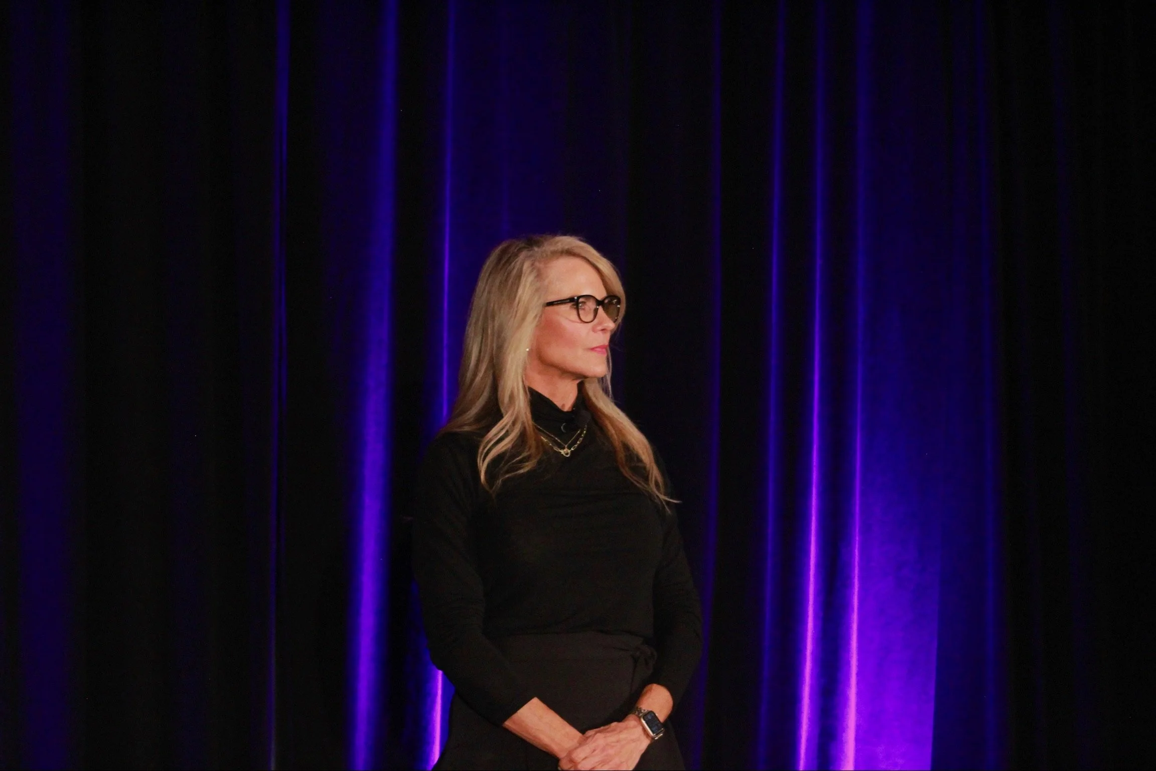 A woman with long blonde hair, wearing glasses, a black turtleneck, and gold jewelry, standing in front of dark curtains illuminated with purple light.