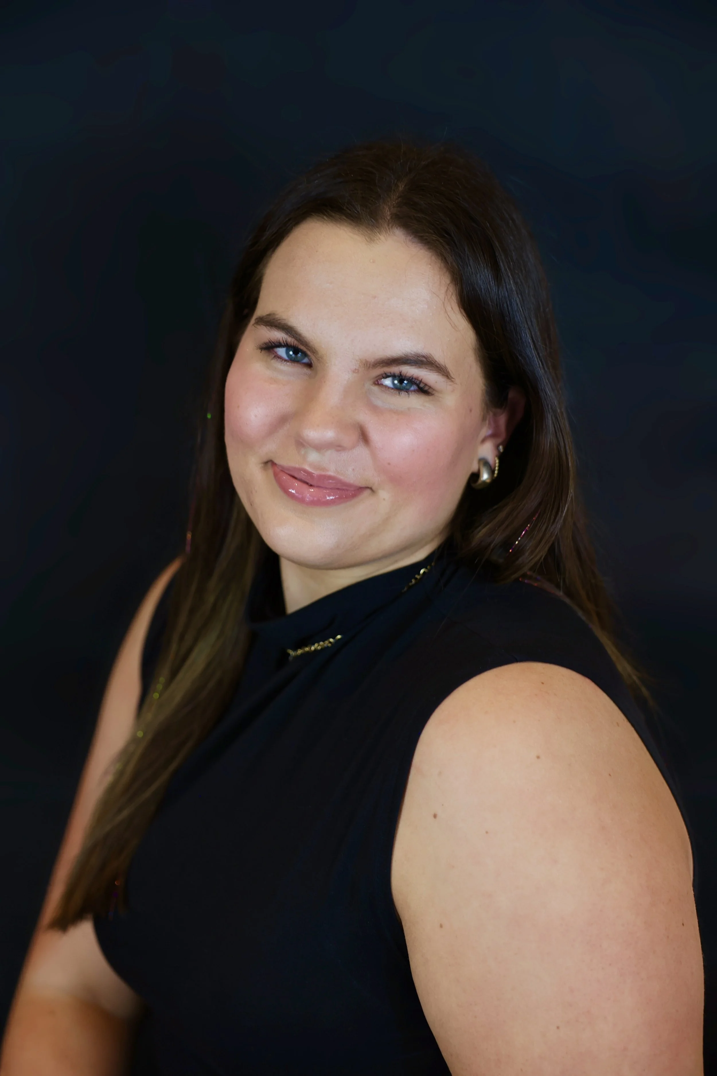 Close-up portrait of a woman with long brown hair, blue eyes, and wearing a black sleeveless top, smiling against a dark background.