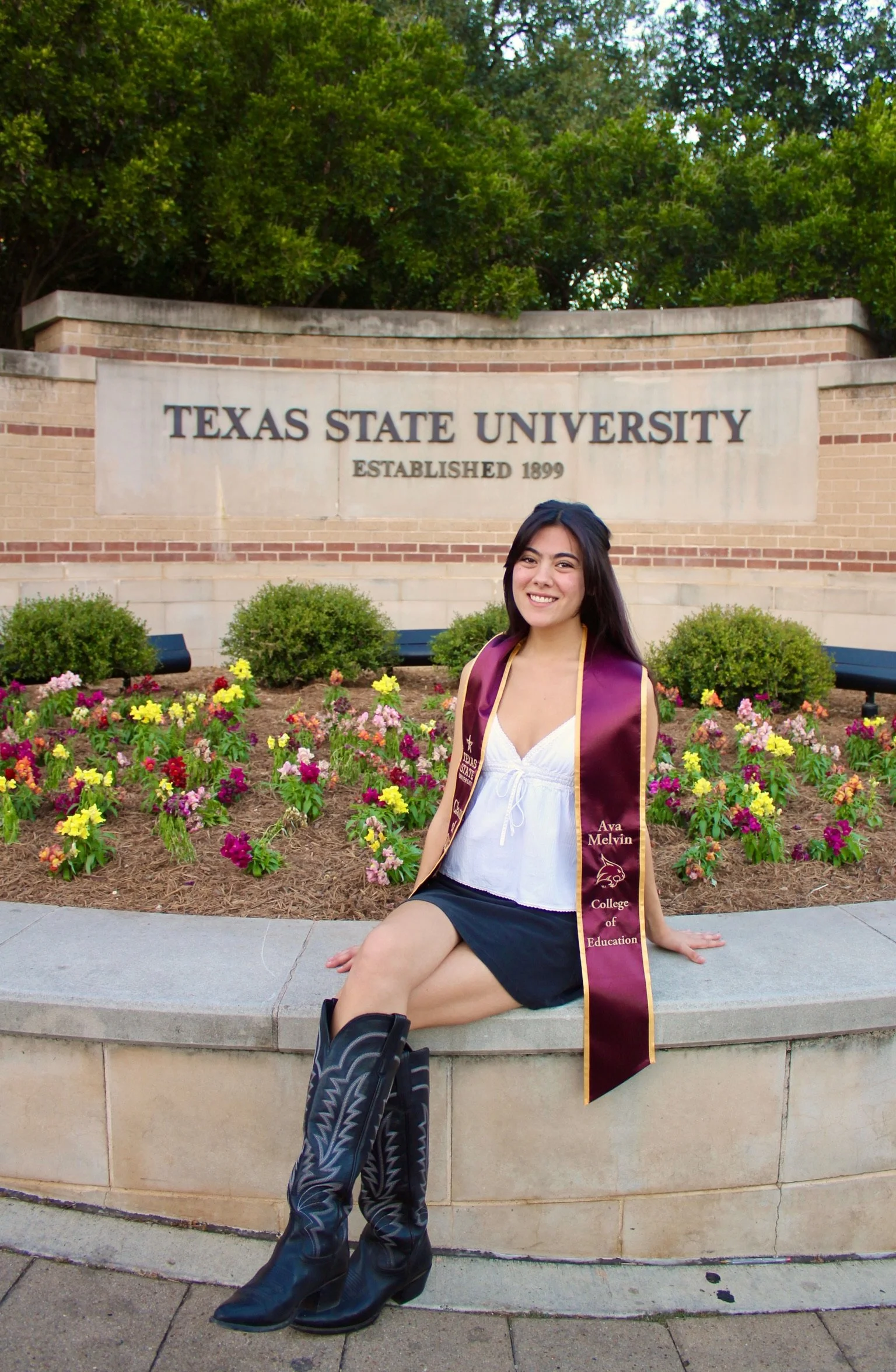 A young woman in a white blouse, black skirt, and boots sitting on a stone ledge in front of a flower garden with orange, pink, and purple flowers, and a sign that reads 'TEXAS STATE UNIVERSITY ESTABLISHED 1899'. She is wearing a maroon and gold stole with her name 'Ava Melvin' and 'College of Education' embroidered on it, and is smiling.