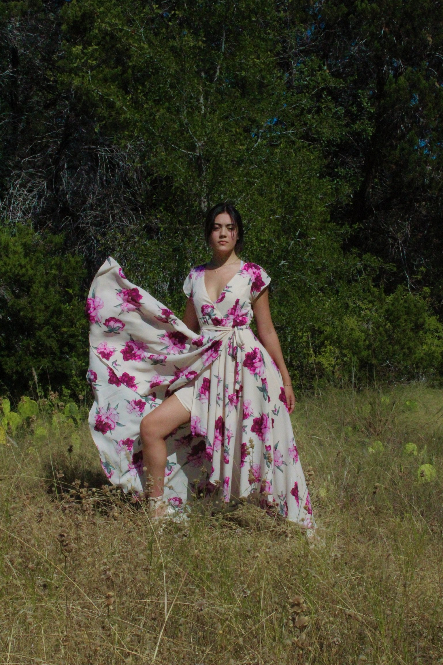 A woman in a floral dress, standing in a grassy field with dense green trees in the background, holding part of her dress up.
