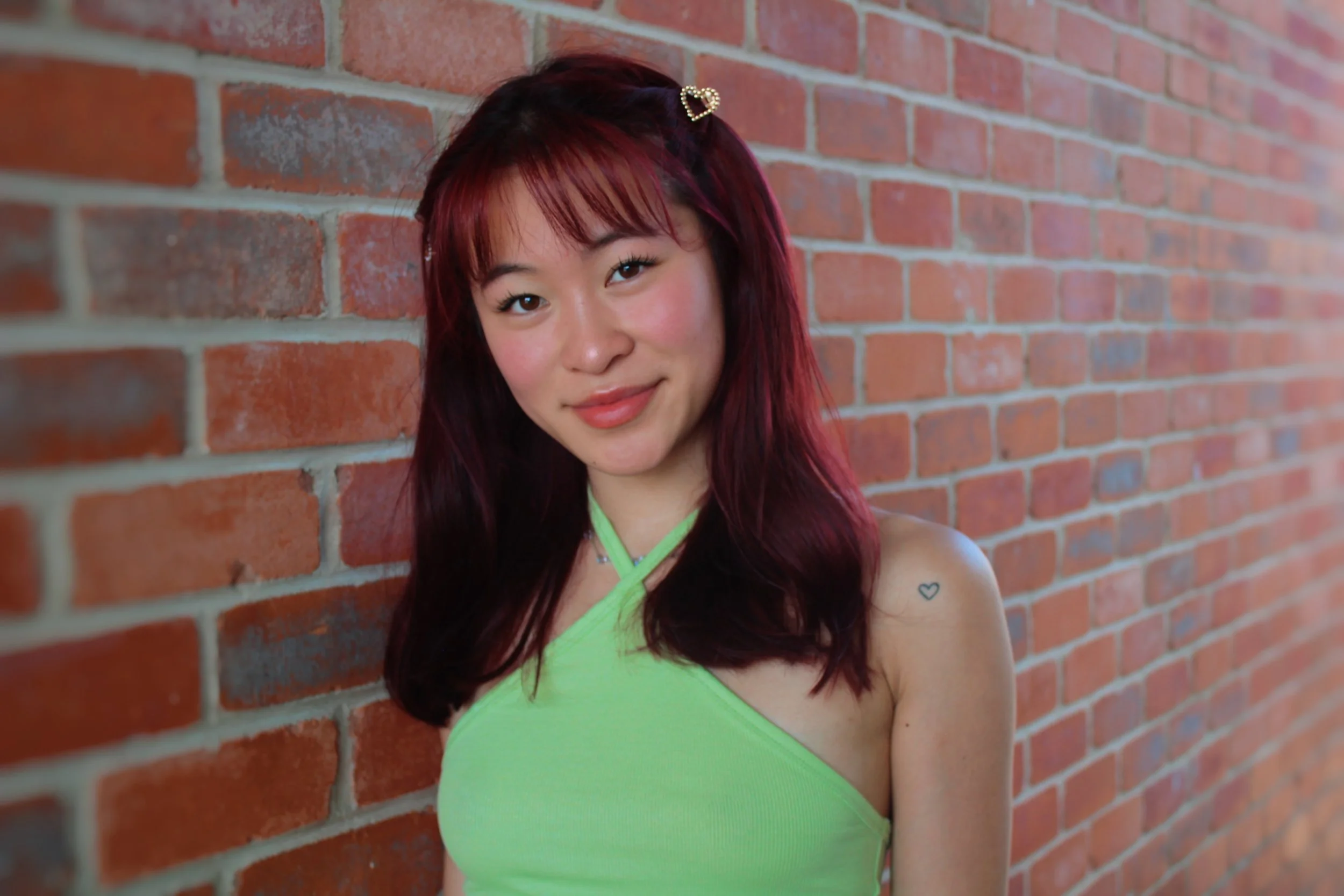 A young woman with red hair and a heart-shaped hair clip Corssely smiling, wearing a neon green top, standing against a brick wall.