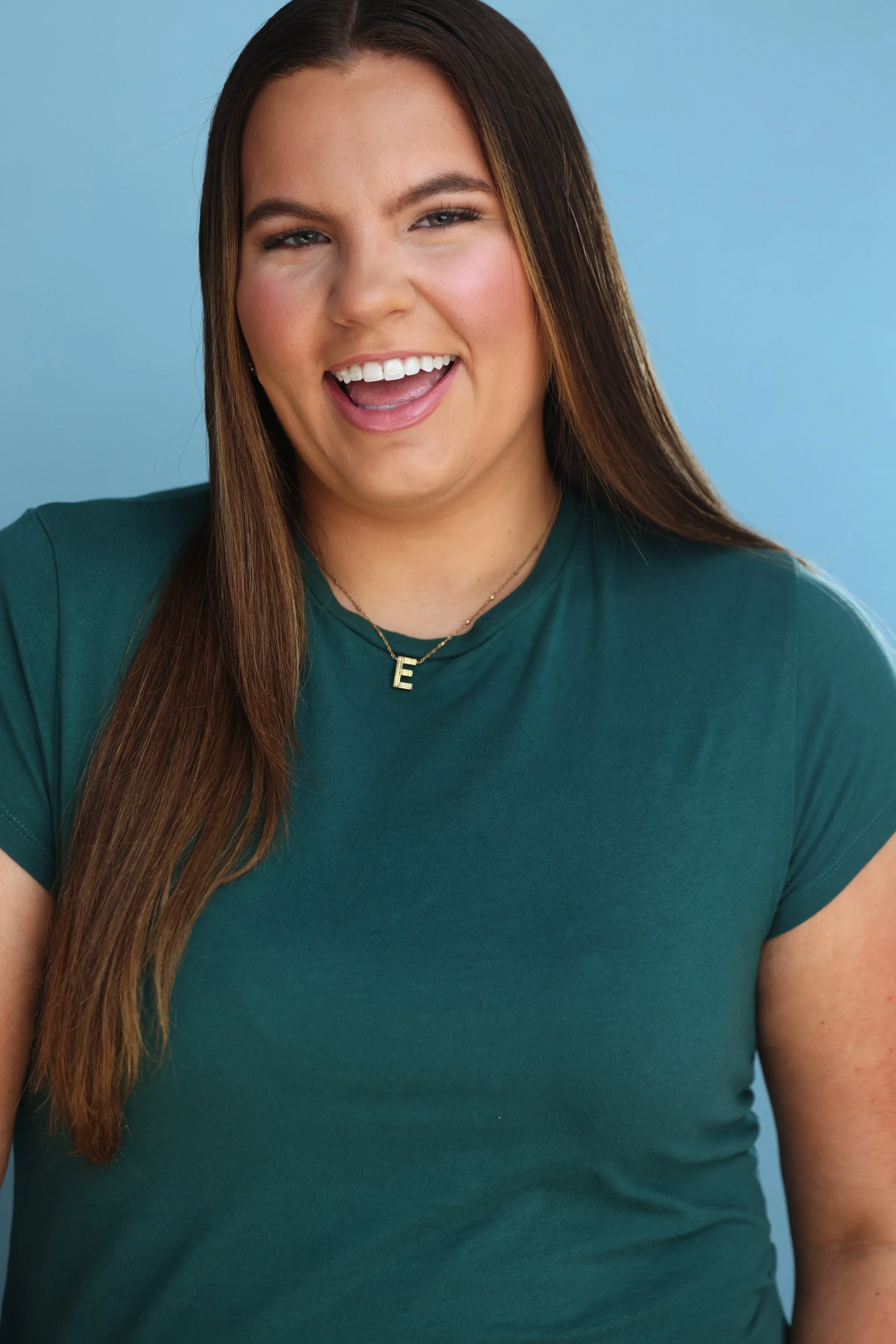 A woman with long brown hair, smiling, wearing a dark green T-shirt and a gold necklace with a letter 'E', against a blue background.