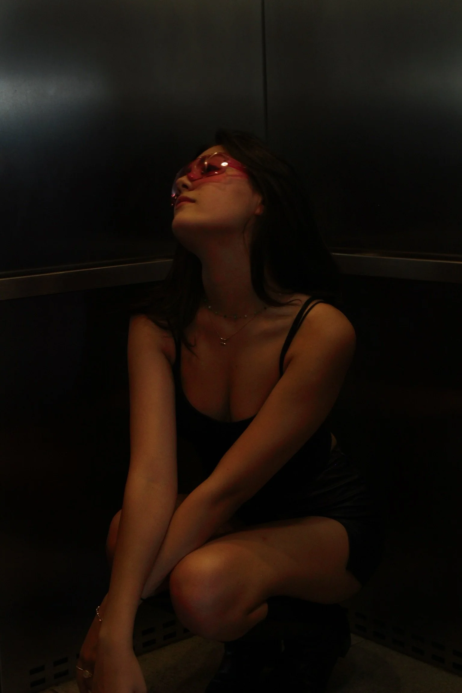 A woman crouching in a dark, enclosed elevator space, wearing sunglasses, a black spaghetti strap top, and jewelry, with her head tilted slightly back.